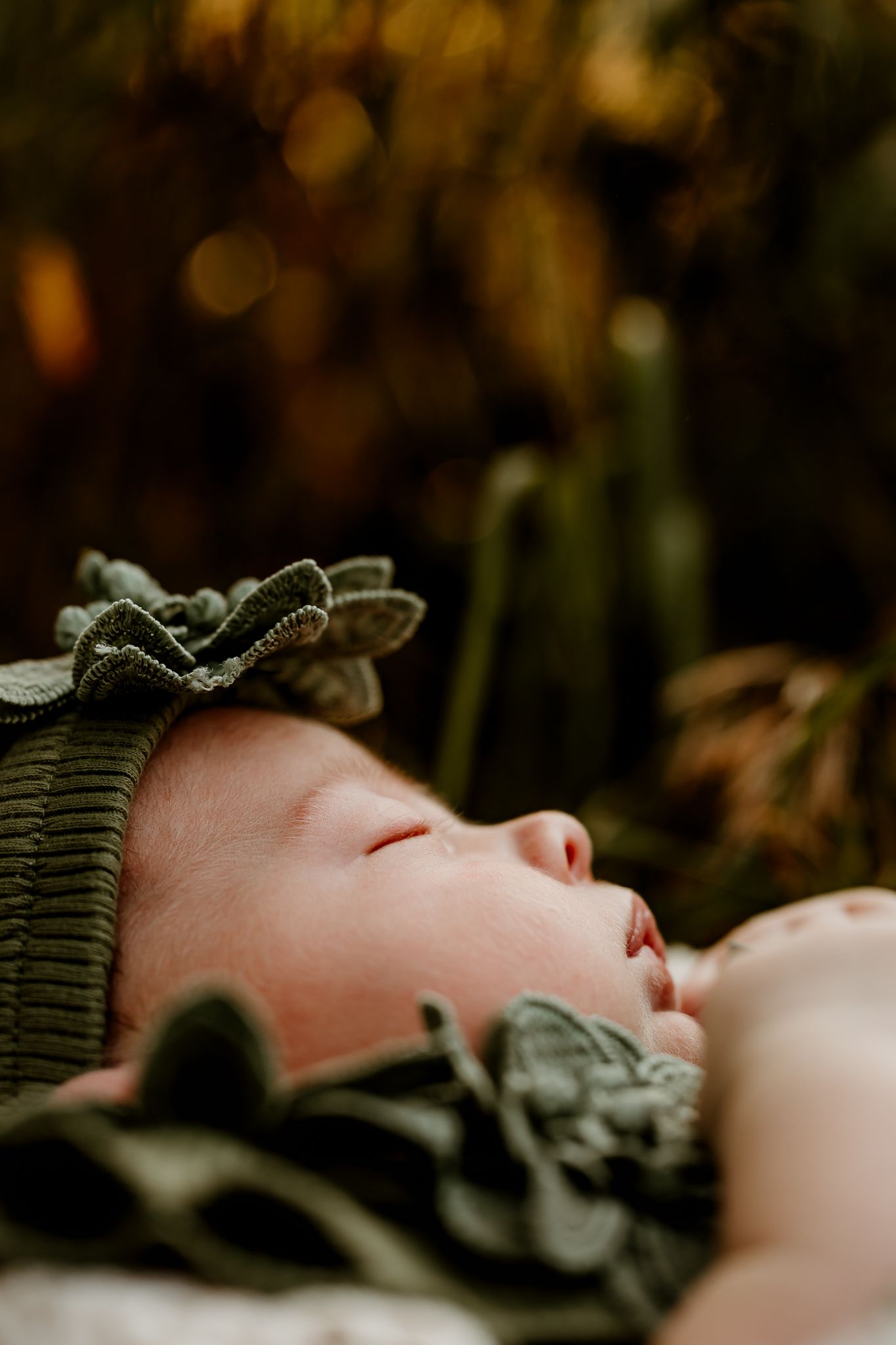 Sleeping newborn baby wearing a green floral bonnet, peacefully resting outdoors with bokeh foliage background.