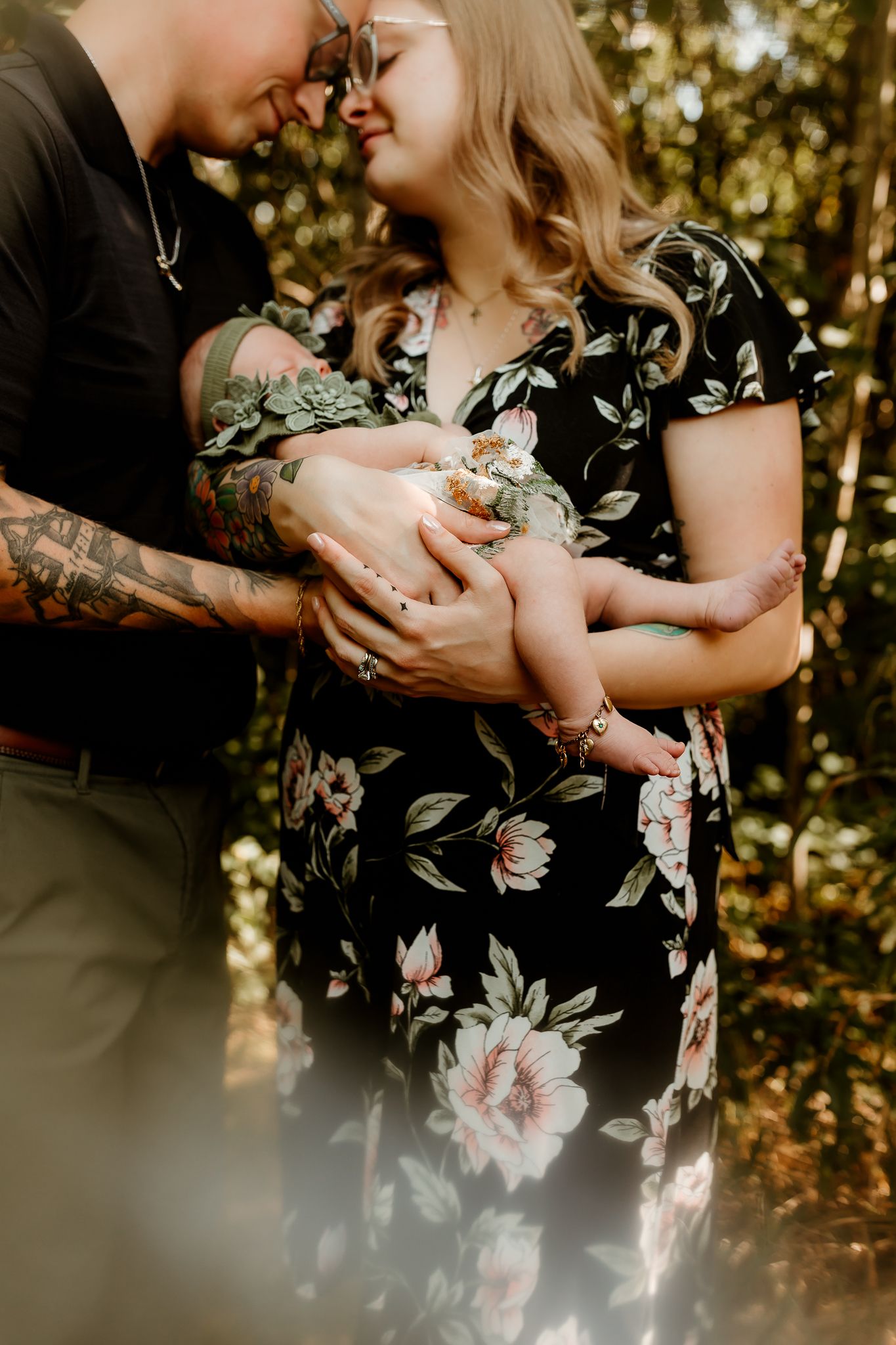 A couple in a forest setting lovingly cradles their newborn baby, the mother wearing a floral dress.