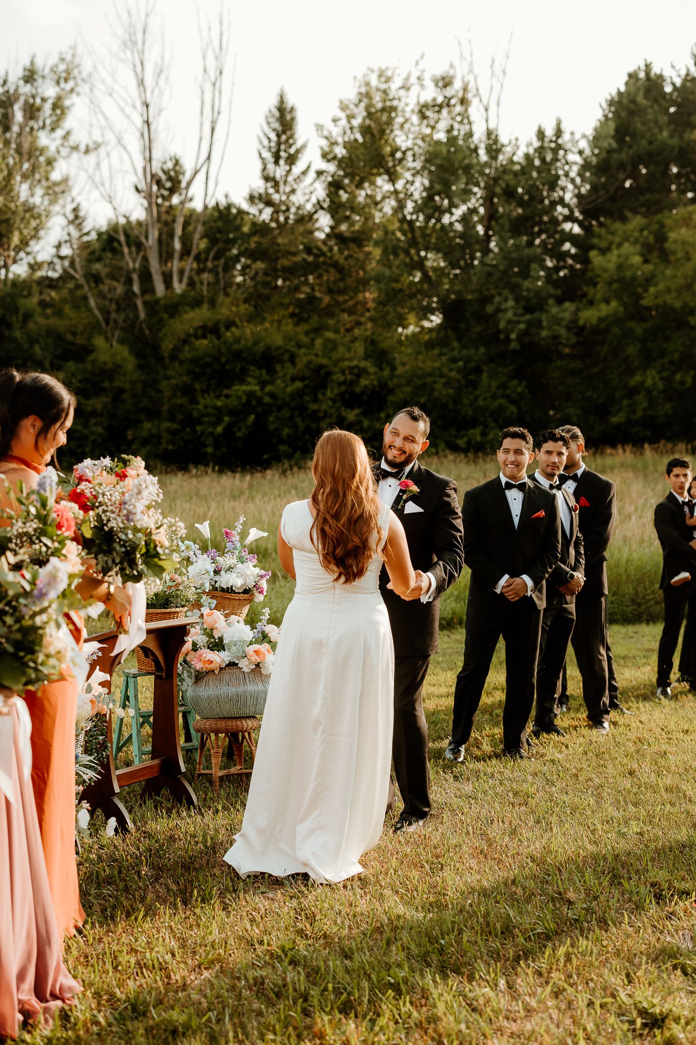 Bride in white gown faces groom in black tuxedo during outdoor wedding ceremony with groomsmen and bridesmaids.