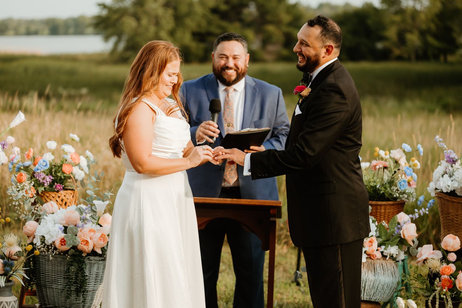 Bride placing ring on groom's finger during outdoor wedding ceremony with officiant and floral arrangements.