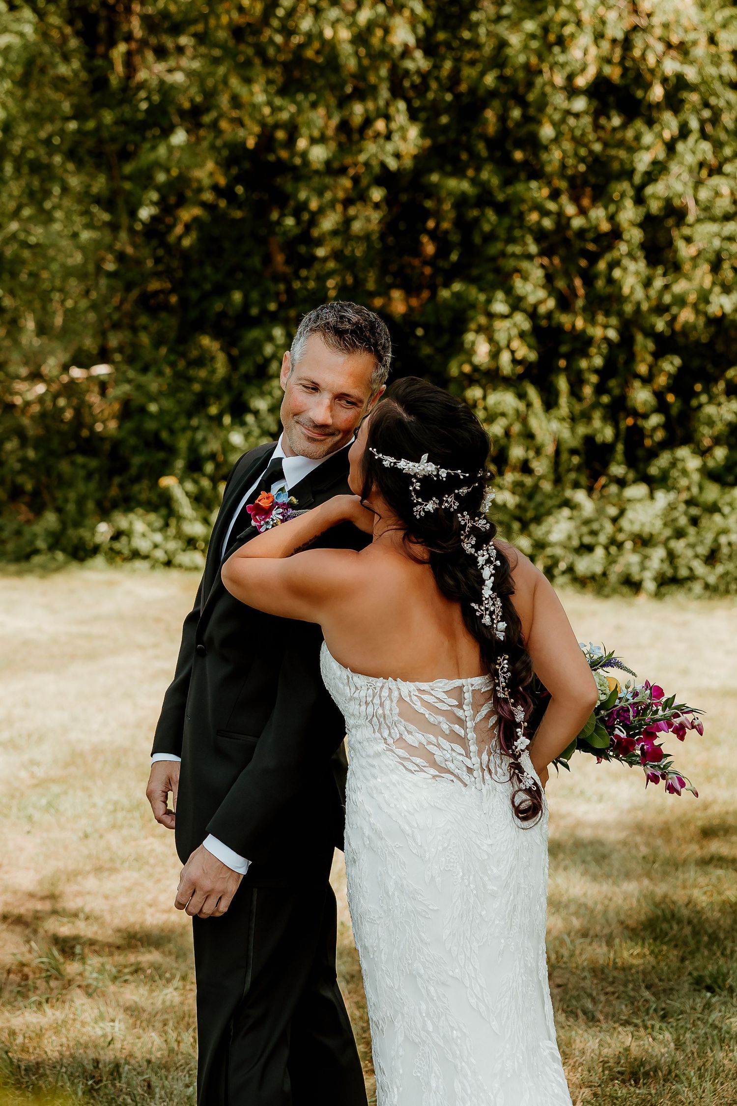 Bride in lace gown holding colorful bouquet embraces groom in black tuxedo during outdoor wedding first look.