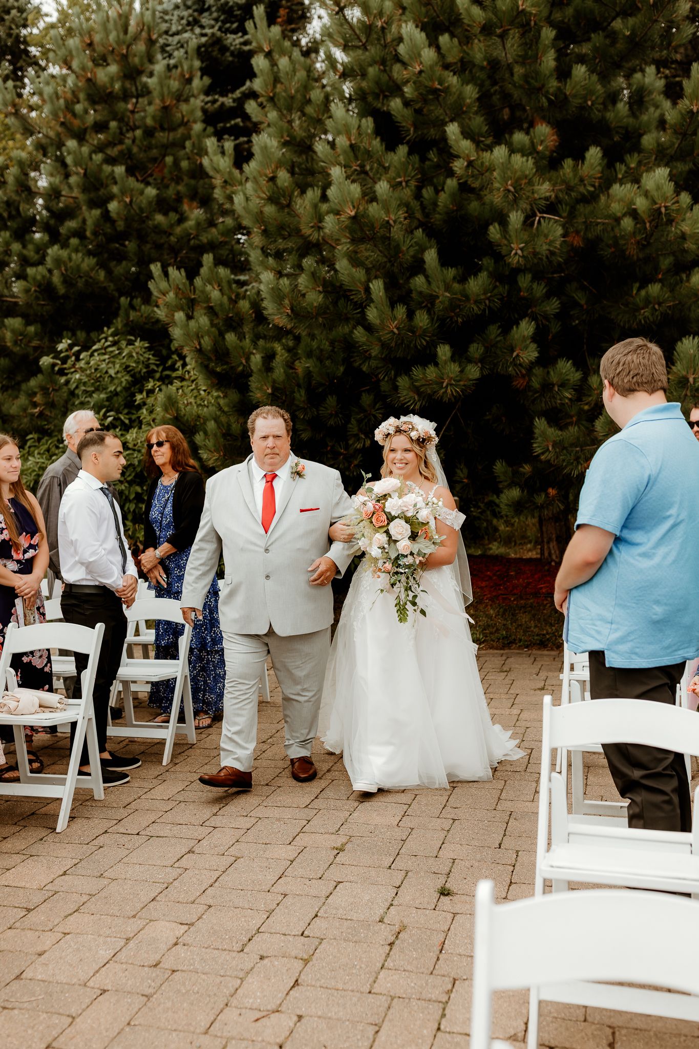 Bride in floral crown walks down outdoor aisle with escort, surrounded by guests and lush pine trees.