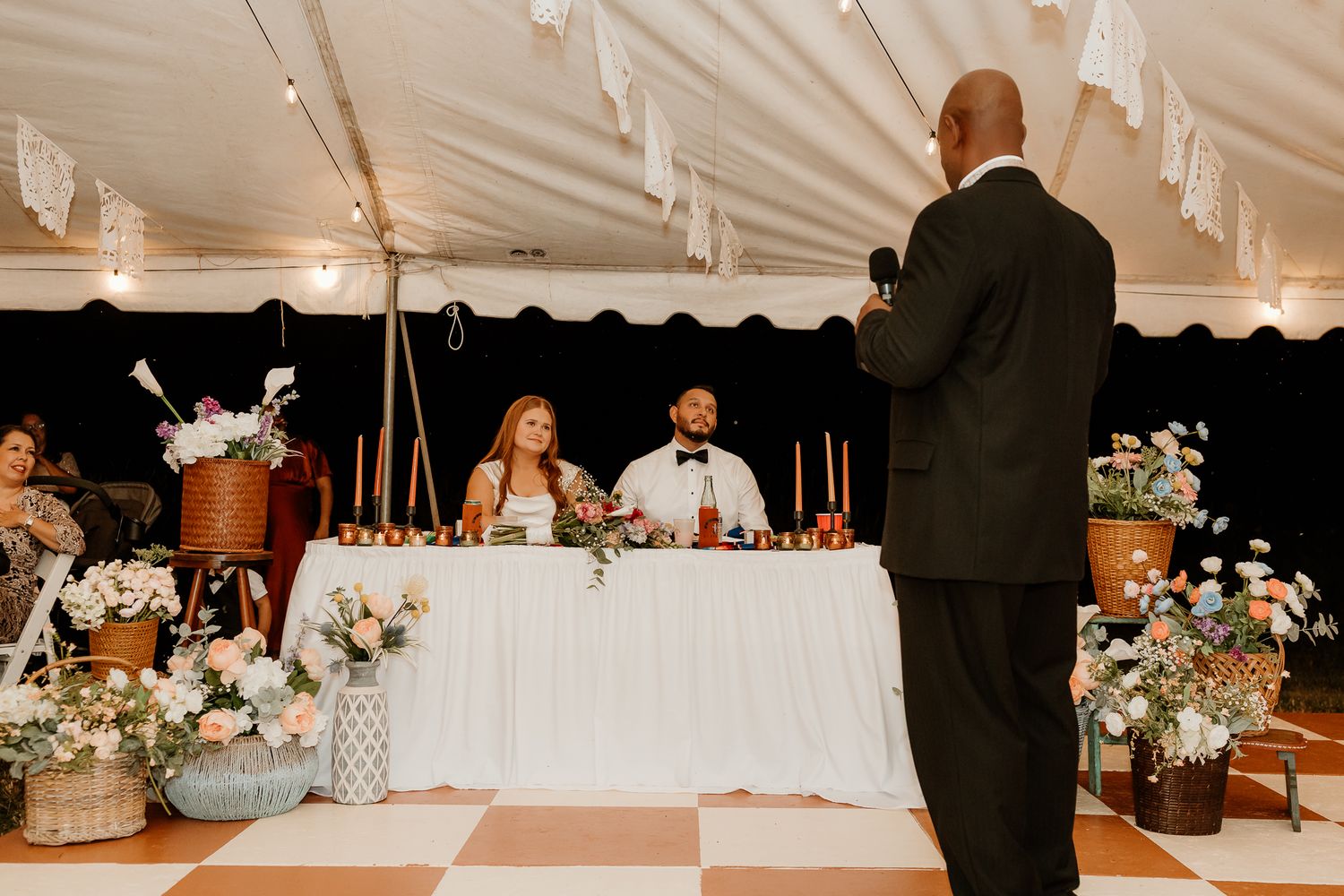 Groom and bride seated at sweetheart table while a man gives a wedding speech inside a tent reception.