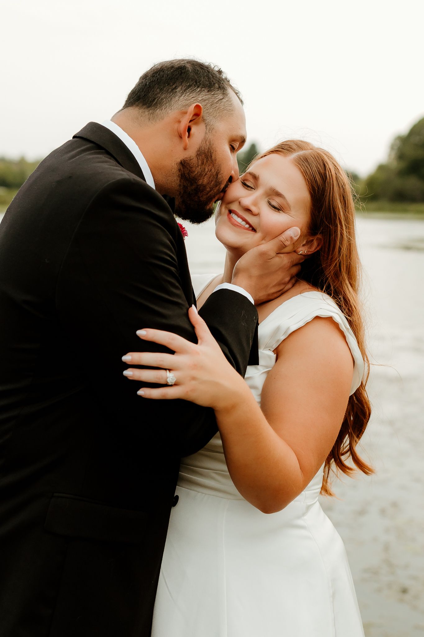 A groom in a black tuxedo kisses his smiling bride in a white dress on the cheek near a serene waterfront.