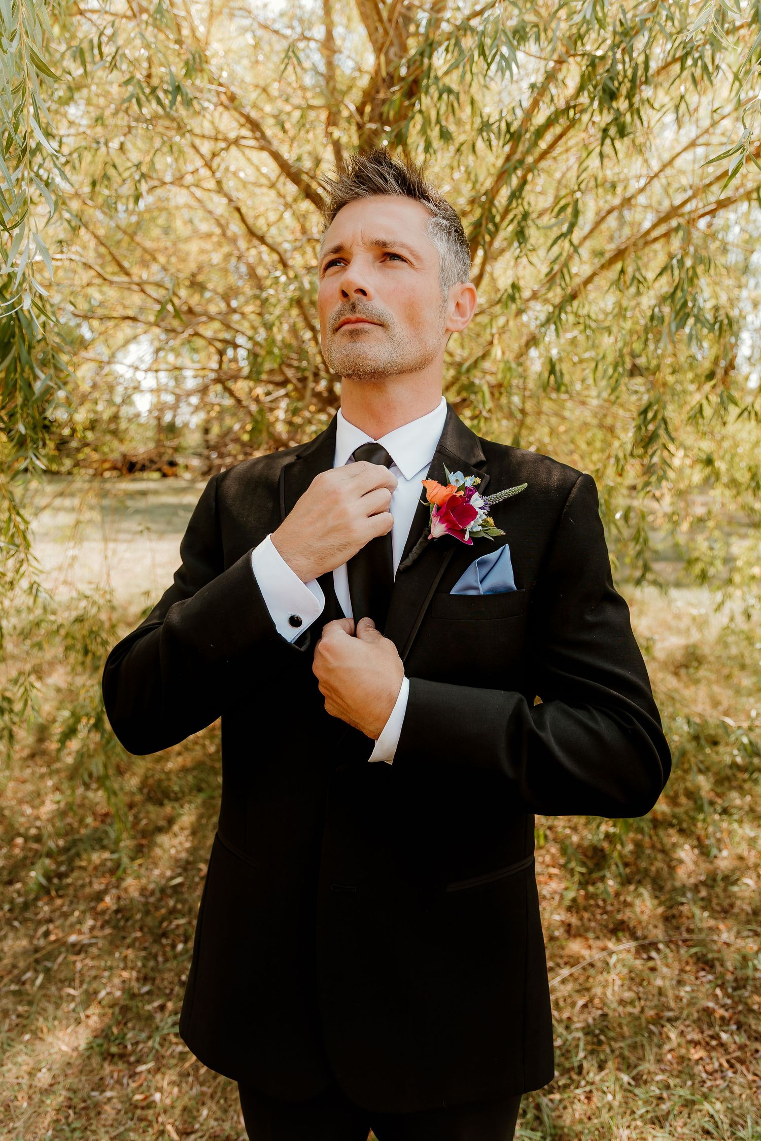 A groom in a black tuxedo adjusts his tie outdoors, wearing a colorful boutonniere and blue pocket square.