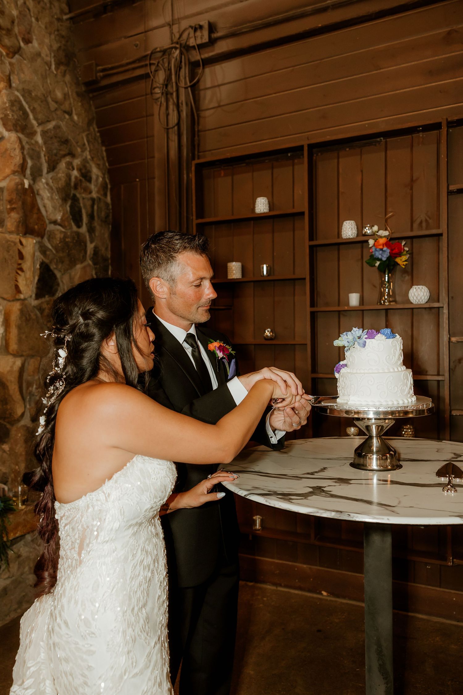 Bride and groom cutting their white wedding cake together in a rustic wood-paneled venue with stone fireplace.
