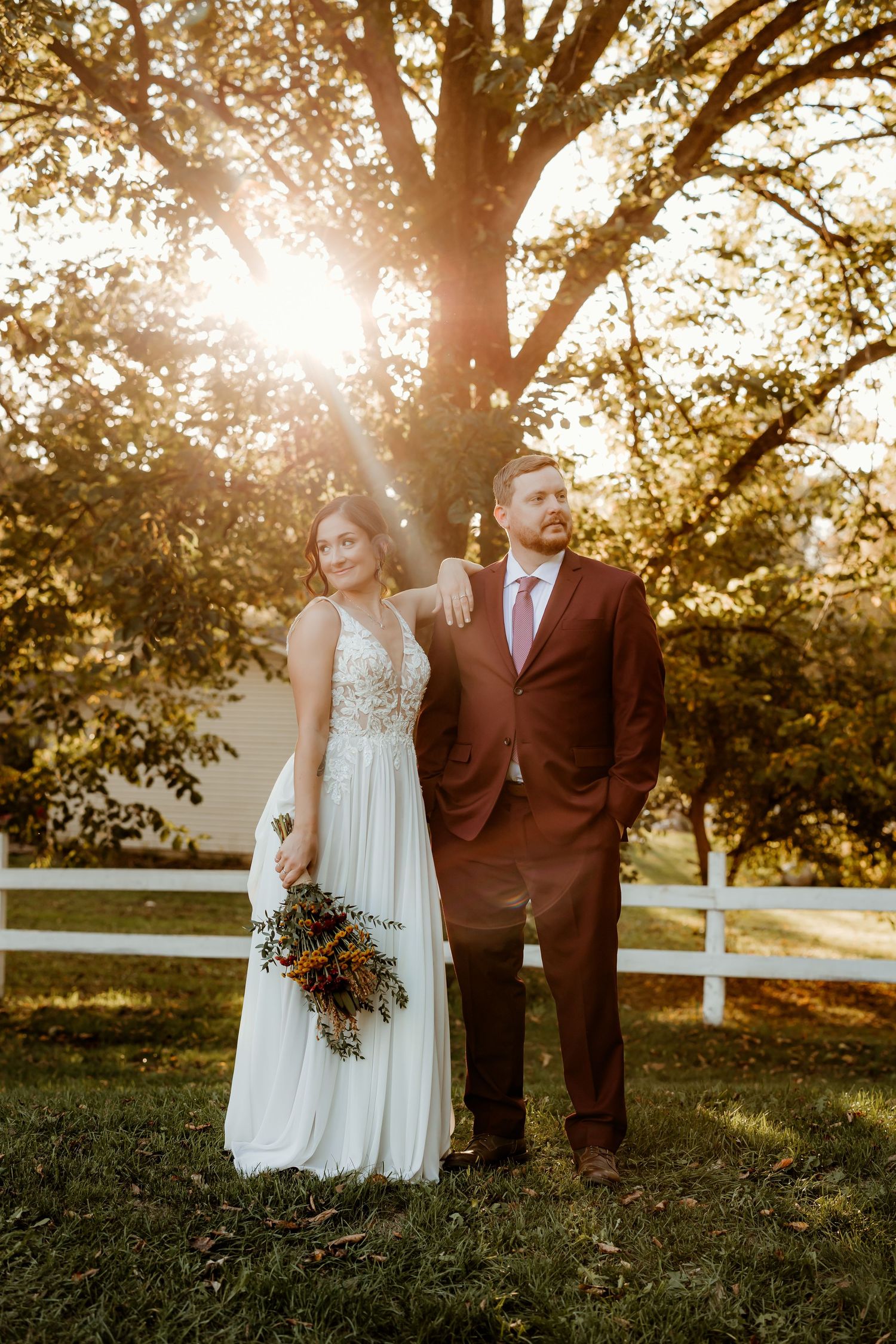 Bride in a lace wedding gown holding an autumn bouquet stands beside a groom in a burgundy suit under golden sunlit trees.