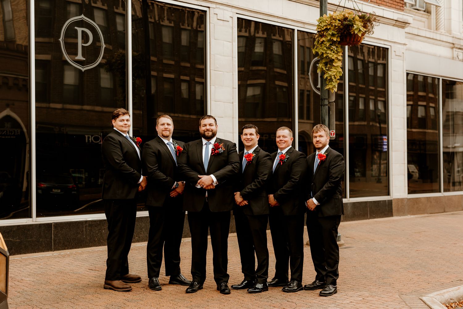 Six groomsmen in black suits with red boutonnieres stand together on a brick sidewalk outside a downtown building for a wedding party photo.