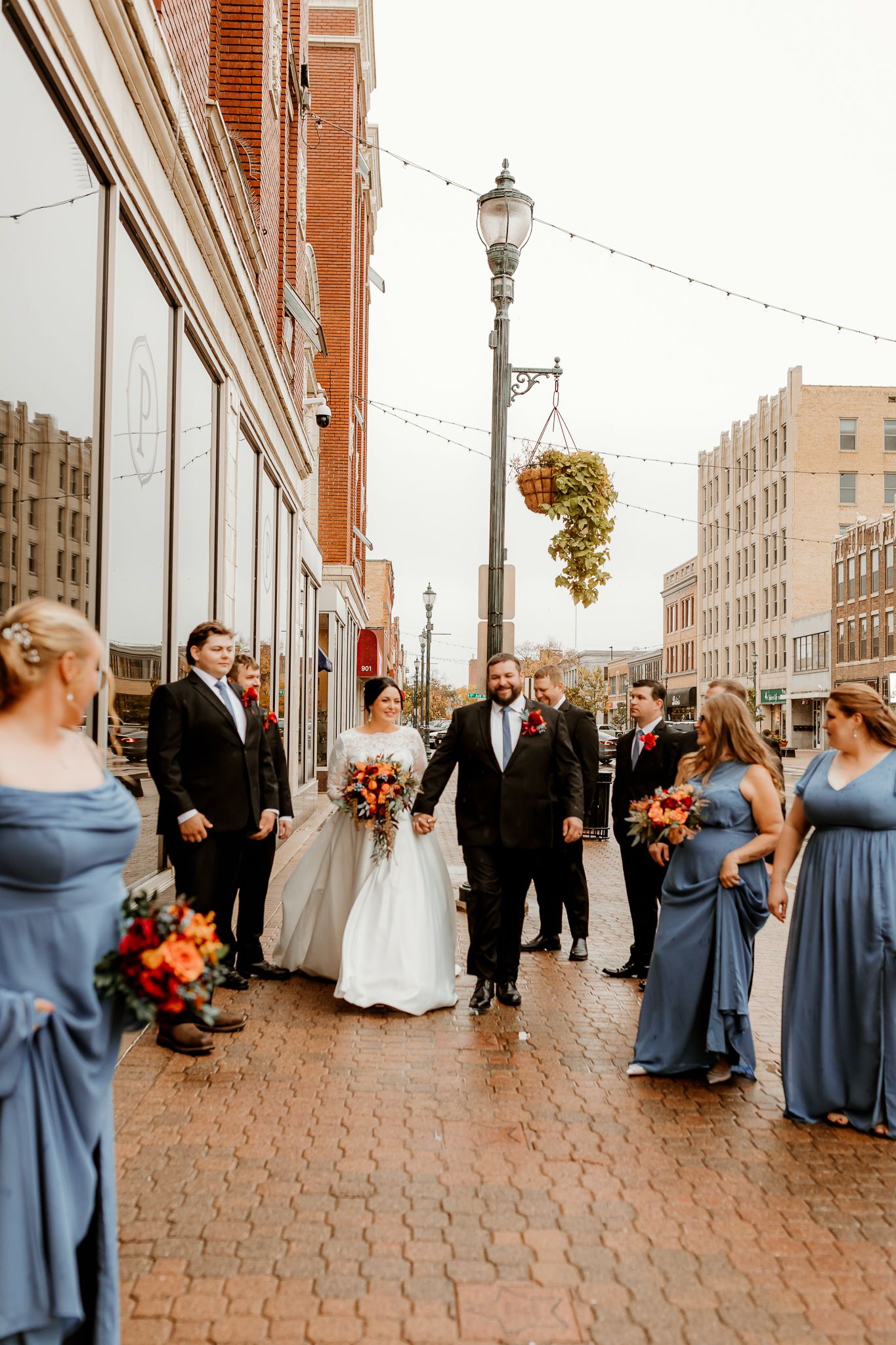 Bride and groom walk arm in arm down a brick city street, flanked by bridesmaids in dusty blue dresses and groomsmen in dark suits.