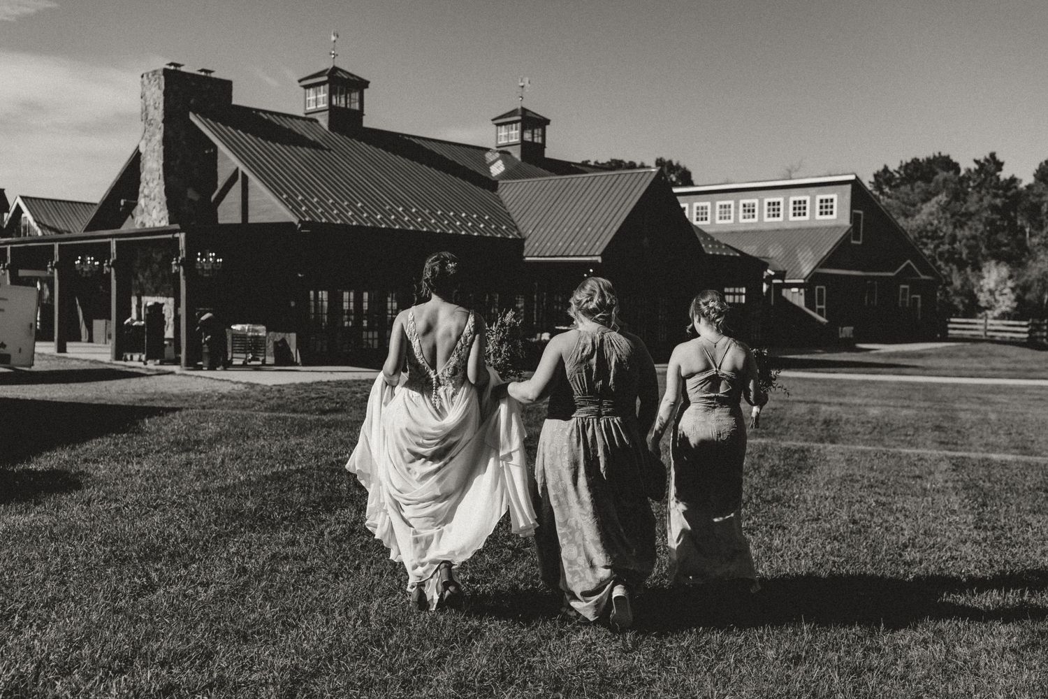 Three women in flowing gowns walk arm in arm across a grassy lawn toward a rustic barn venue in a black and white wedding photo.