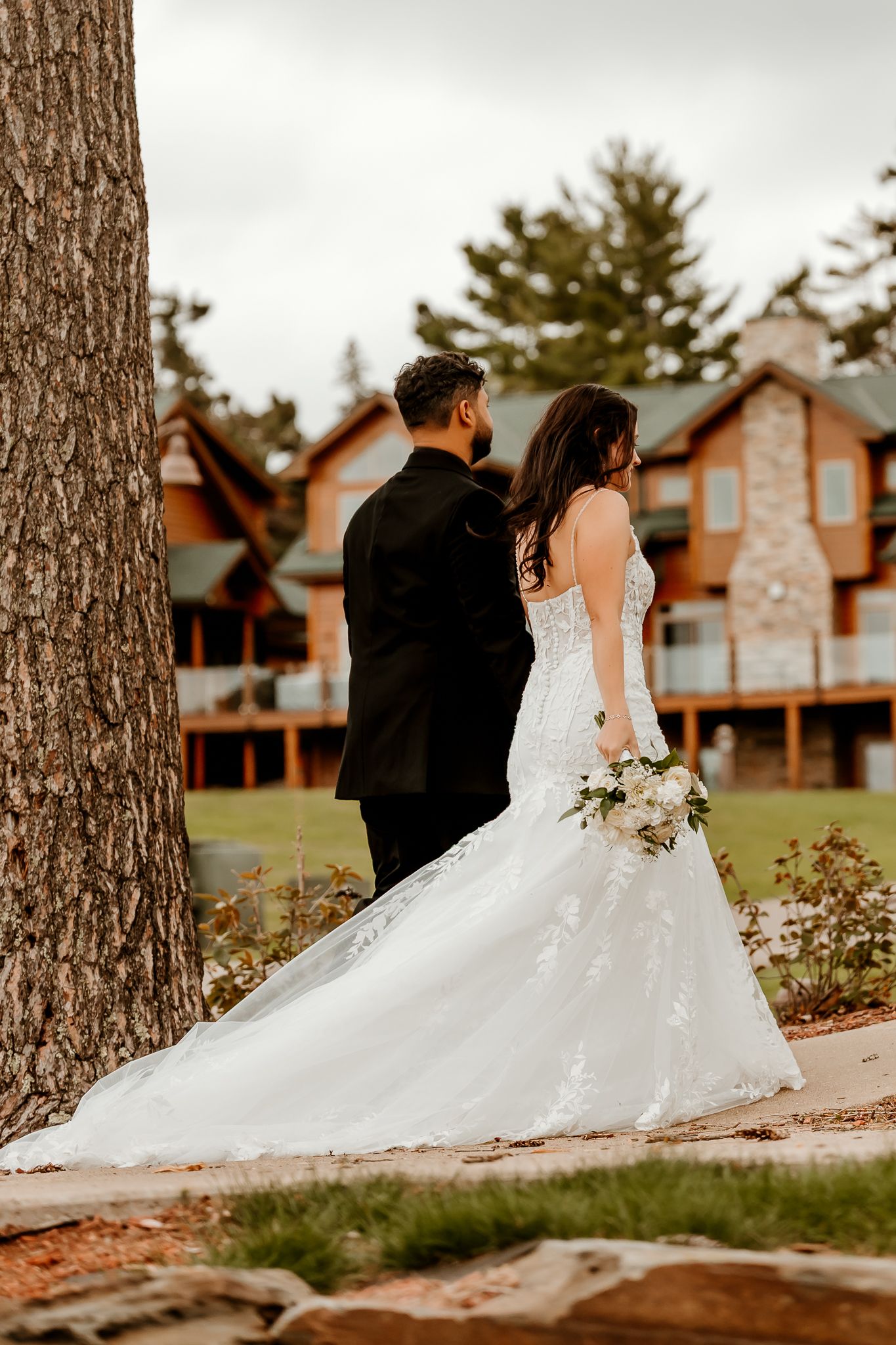 Bride in a lace wedding gown holding a white bouquet stands beside a groom in a black suit near a tree with a wooden lodge behind them.