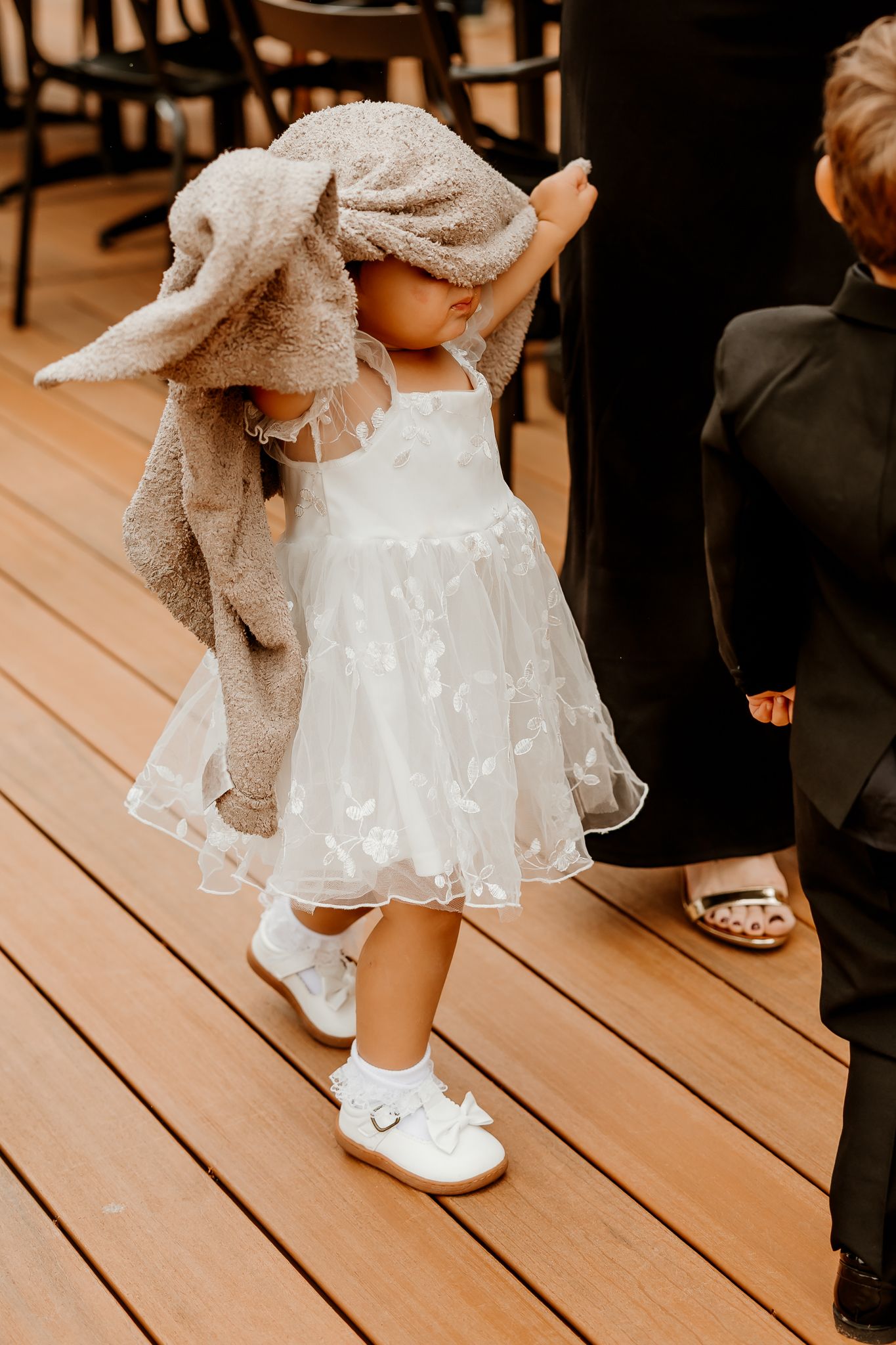 Toddler in a white tulle flower girl dress and white shoes wears a gray sweater over her head on a wooden deck beside a boy in a suit.