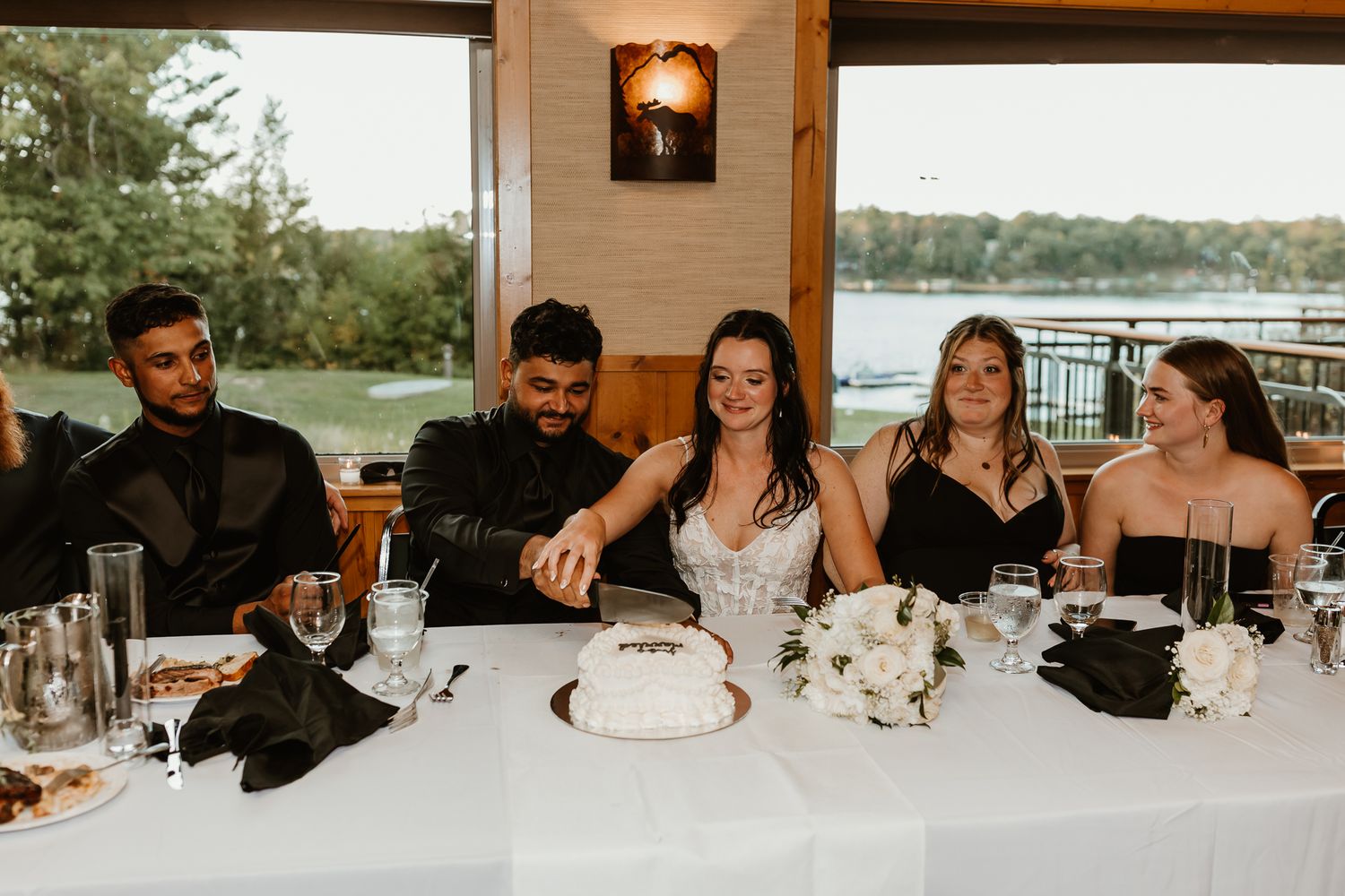Bride and groom cut their wedding cake together at a head table with guests seated beside them and a lake view through the windows.