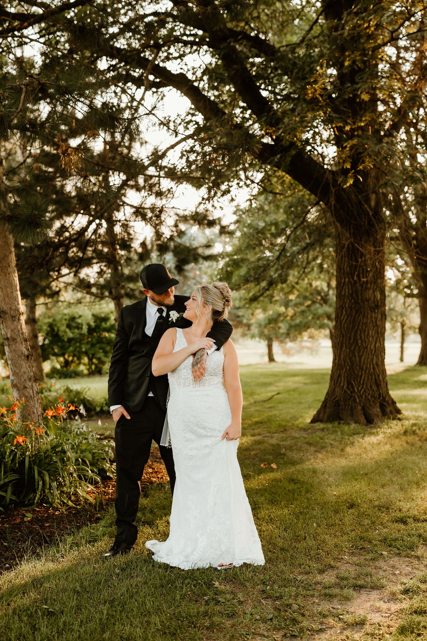 Bride and groom share a romantic moment under large trees in a lush garden setting on their wedding day.