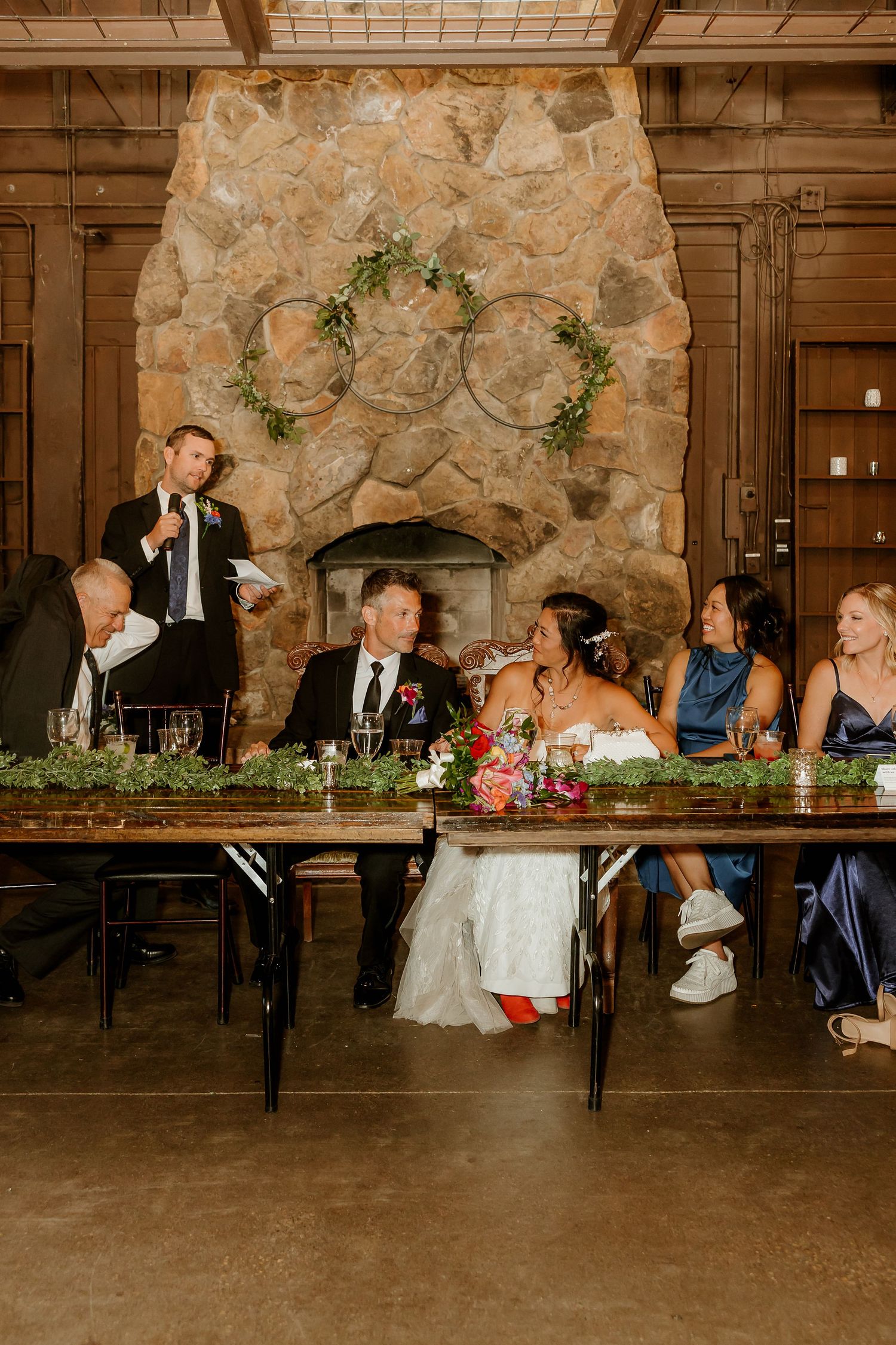 Wedding reception head table with bride and groom seated before a large stone fireplace while a guest delivers a toast.