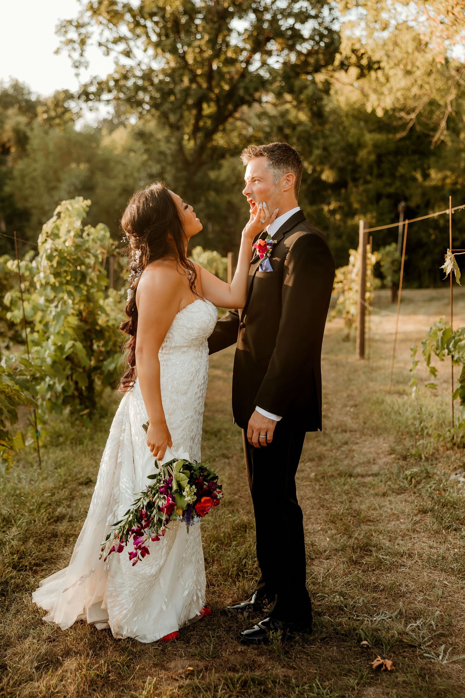 Bride and groom share a tender moment outdoors at golden hour, she in lace gown holding red bouquet, he in black tuxedo.