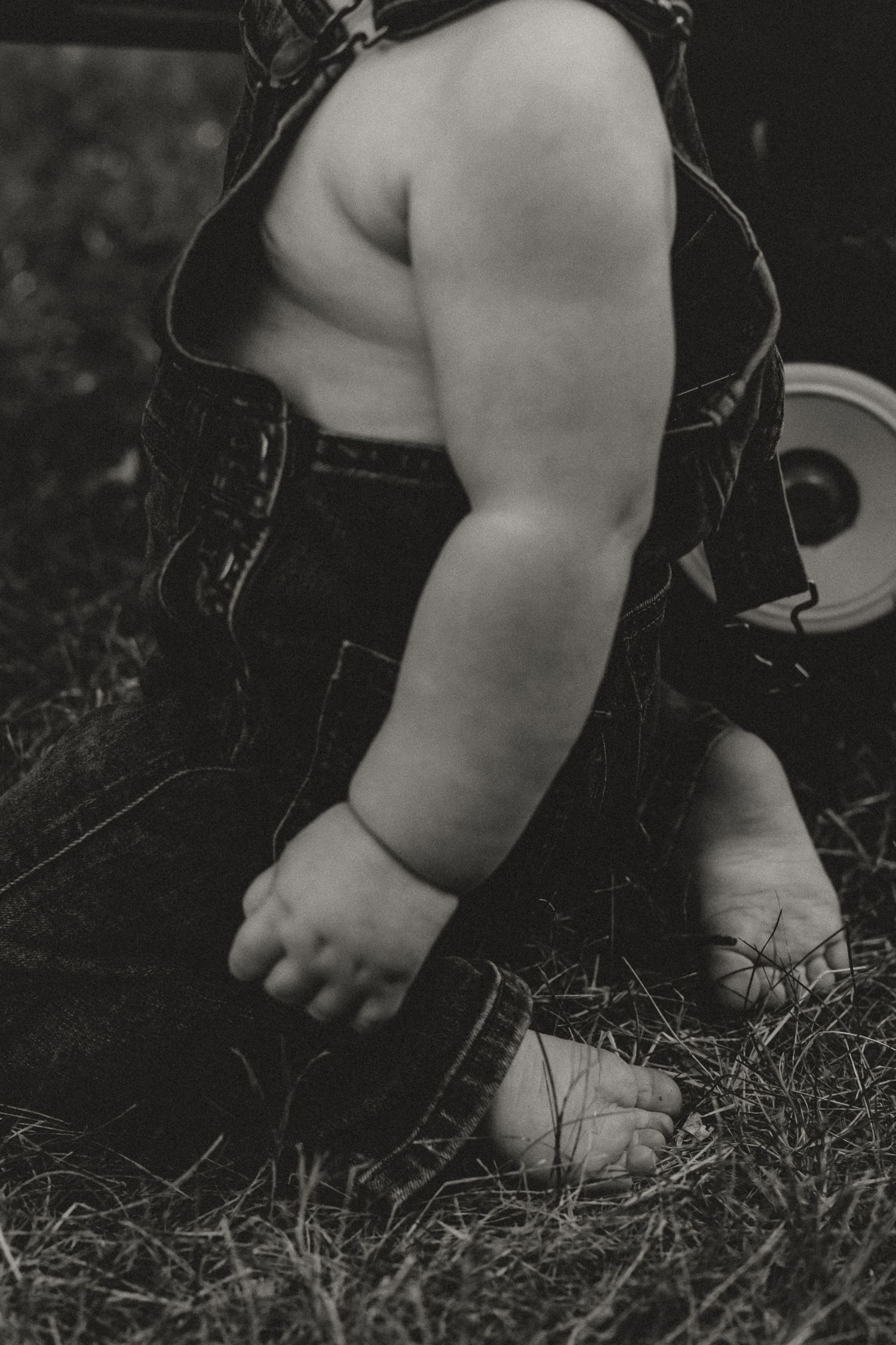 Black and white photo of a chubby toddler in overalls sitting barefoot on grass outdoors.