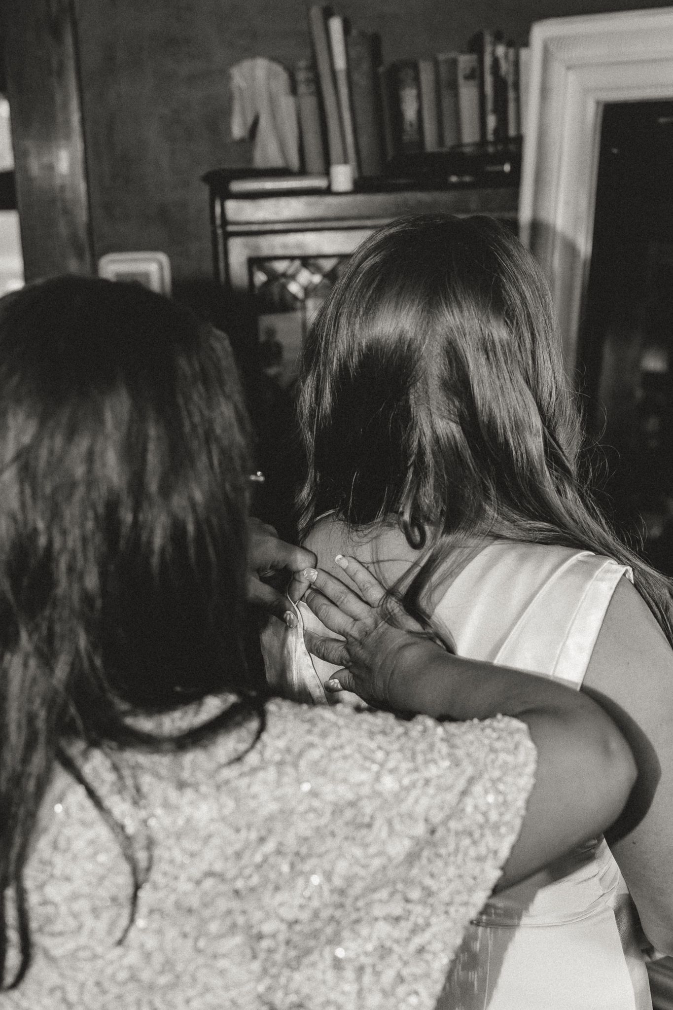 Black and white photo of two girls hugging indoors, one wearing a white dress, with bookshelves in the background.