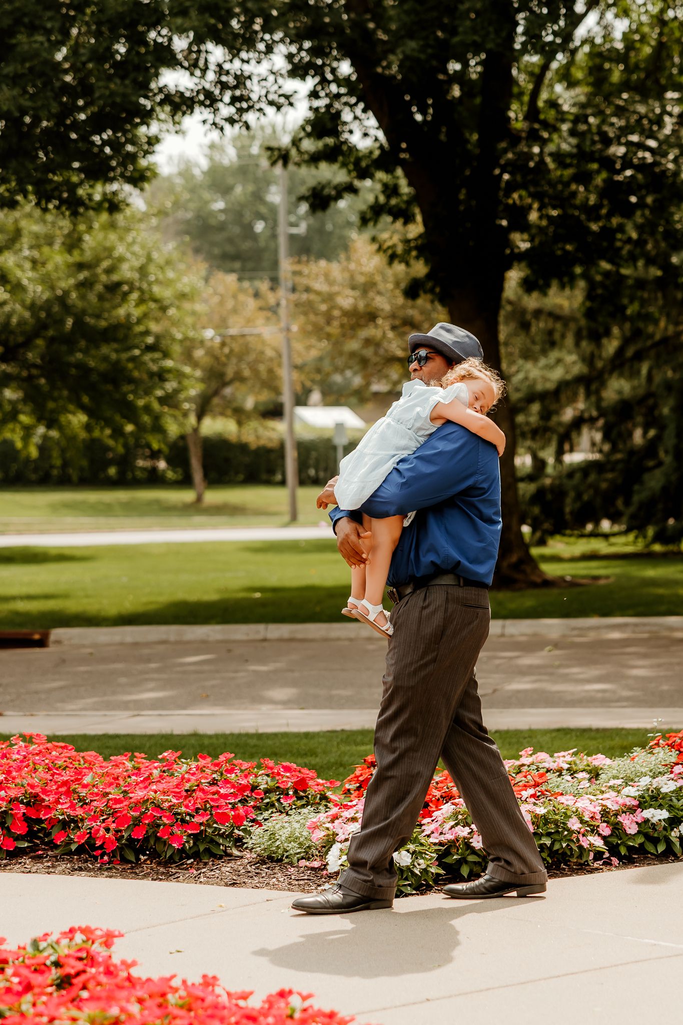 Man in blue shirt and cap carries a young child along a garden path lined with red and pink flowers.