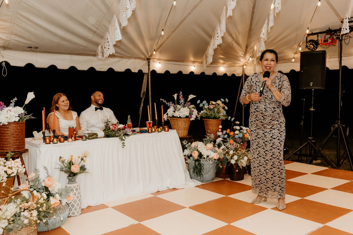 Woman giving a speech at an elegant wedding reception under a tent with floral decorations and checkered dance floor.