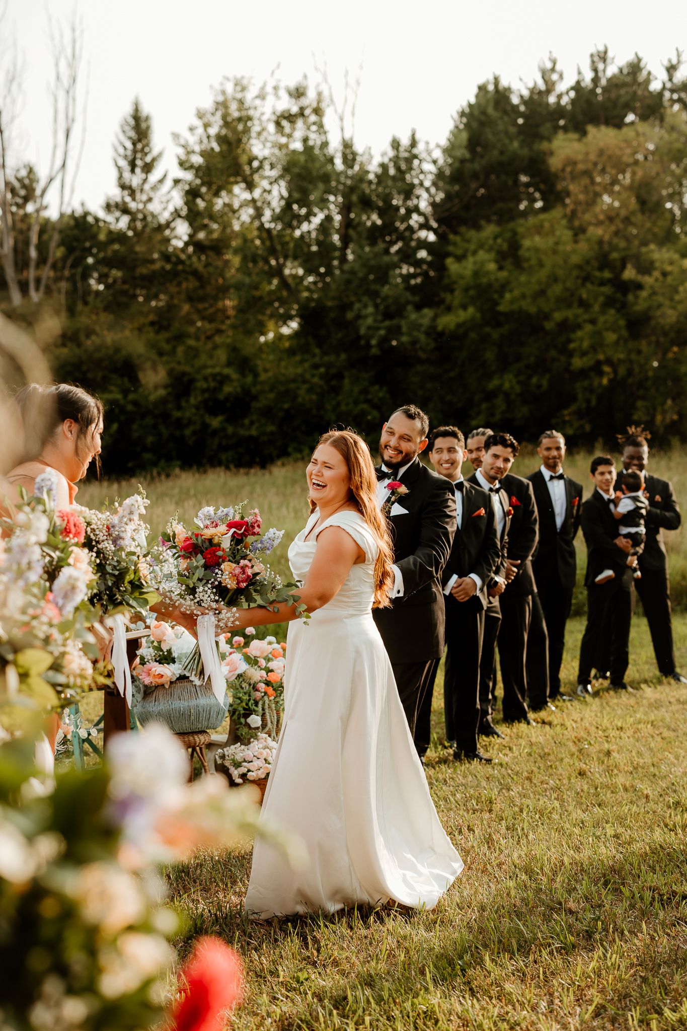 Bride laughing with bouquet during outdoor wedding ceremony, groomsmen in black tuxedos lined up on green lawn.