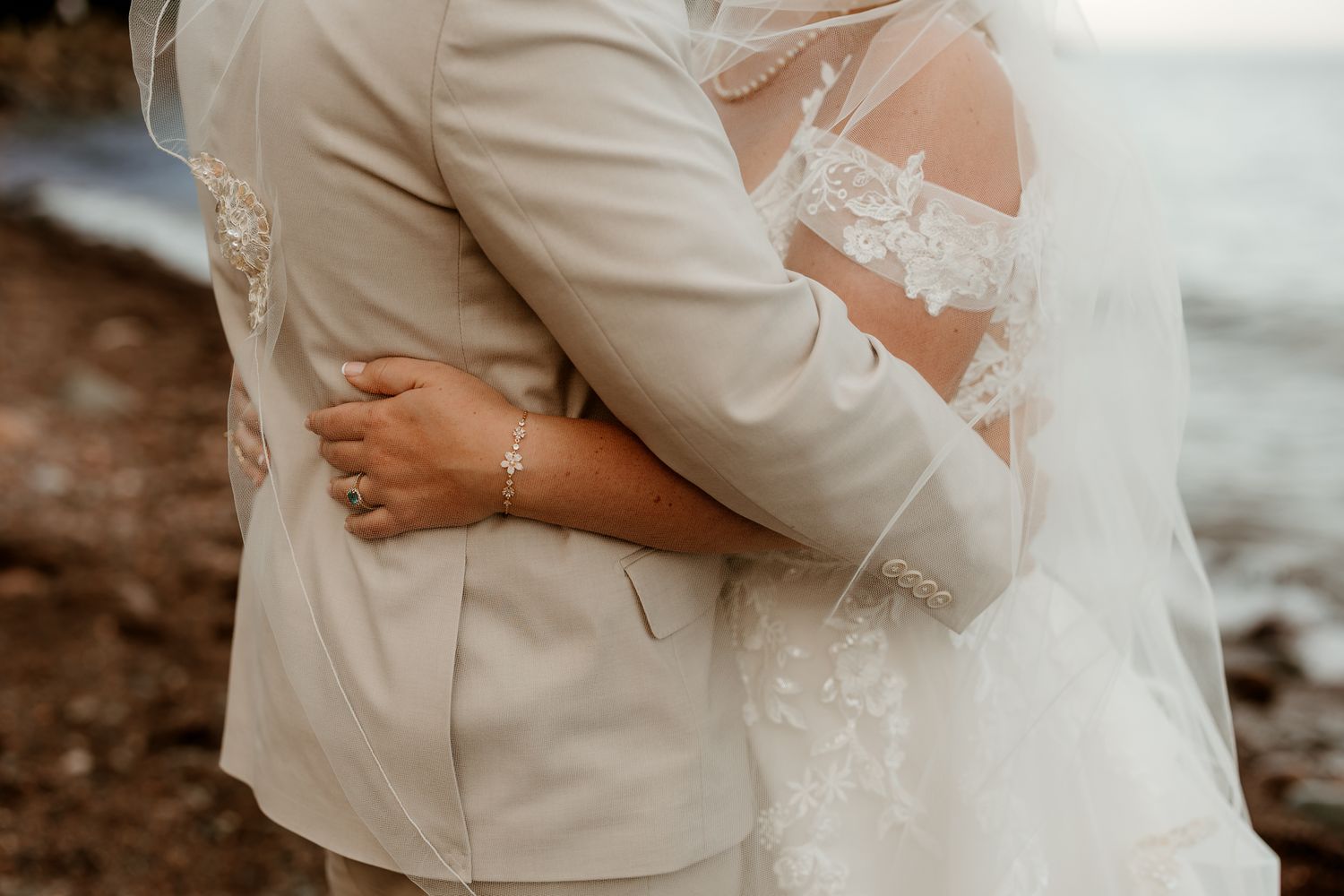 Bride and groom embracing outdoors, wearing lace wedding dress with veil and beige suit near ocean.