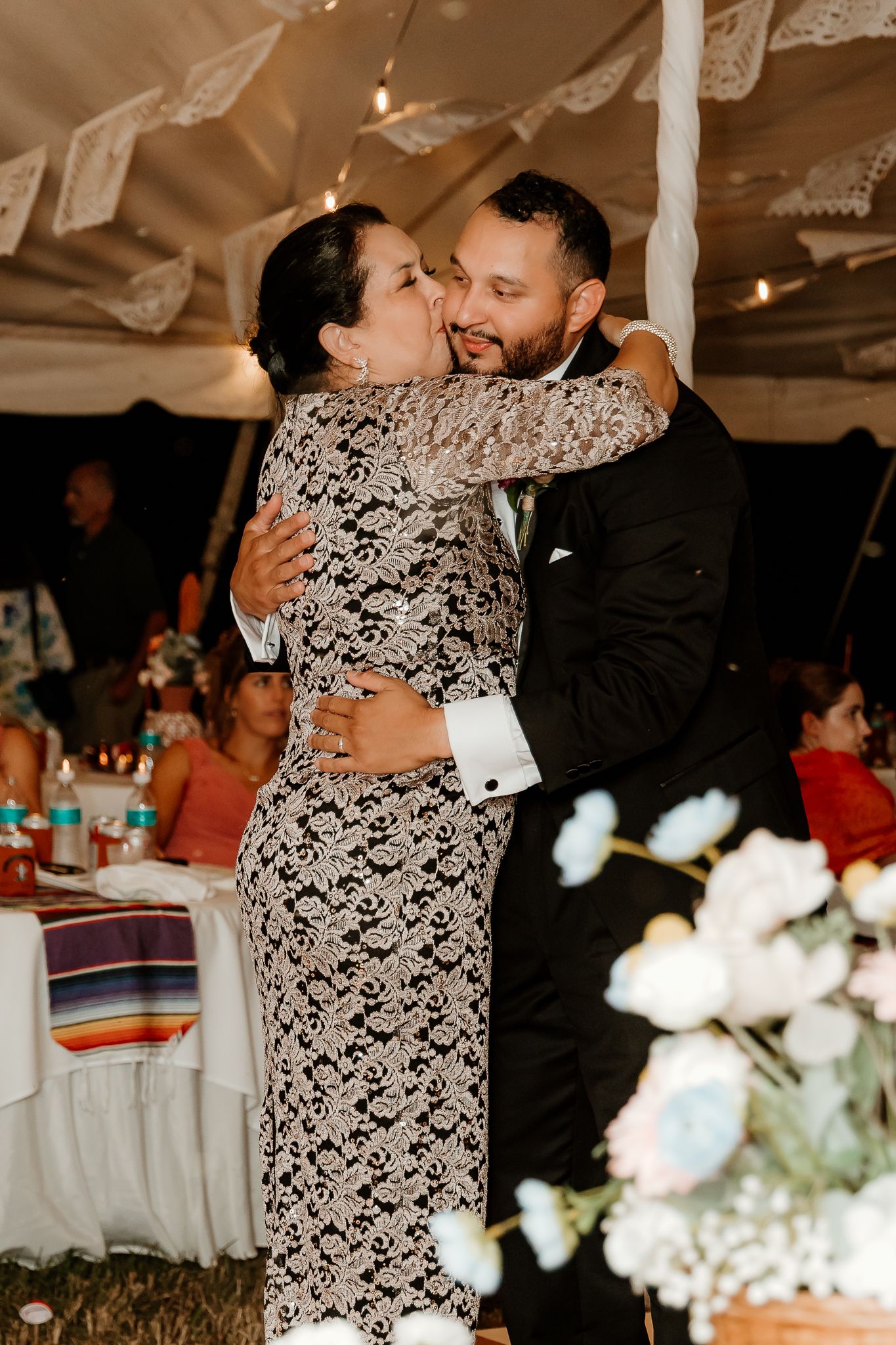 A groom in a black suit shares a tender embrace with a woman in a lace dress at a wedding reception.