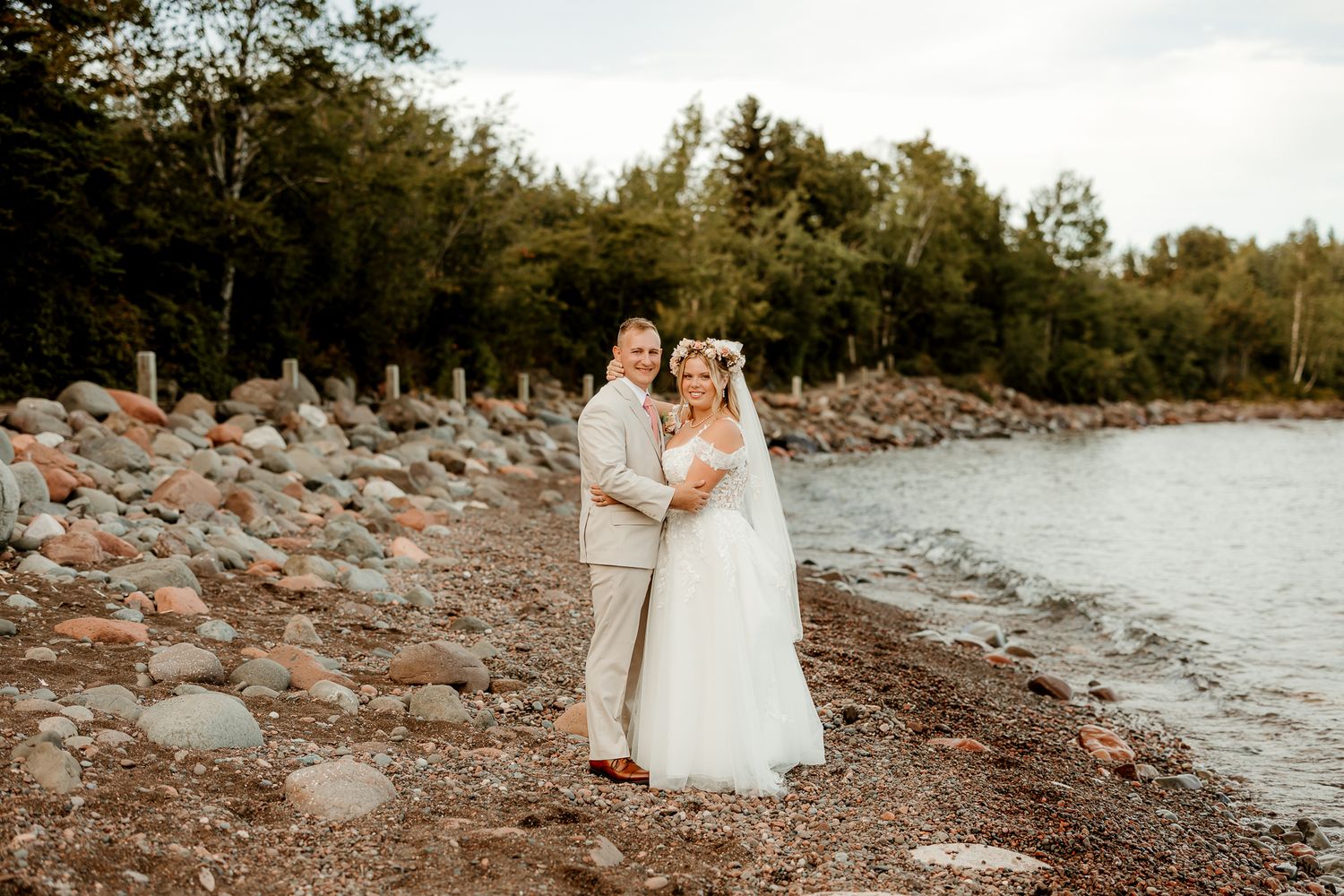 A bride and groom pose together on a rocky lakeside beach surrounded by lush green trees on their wedding day.