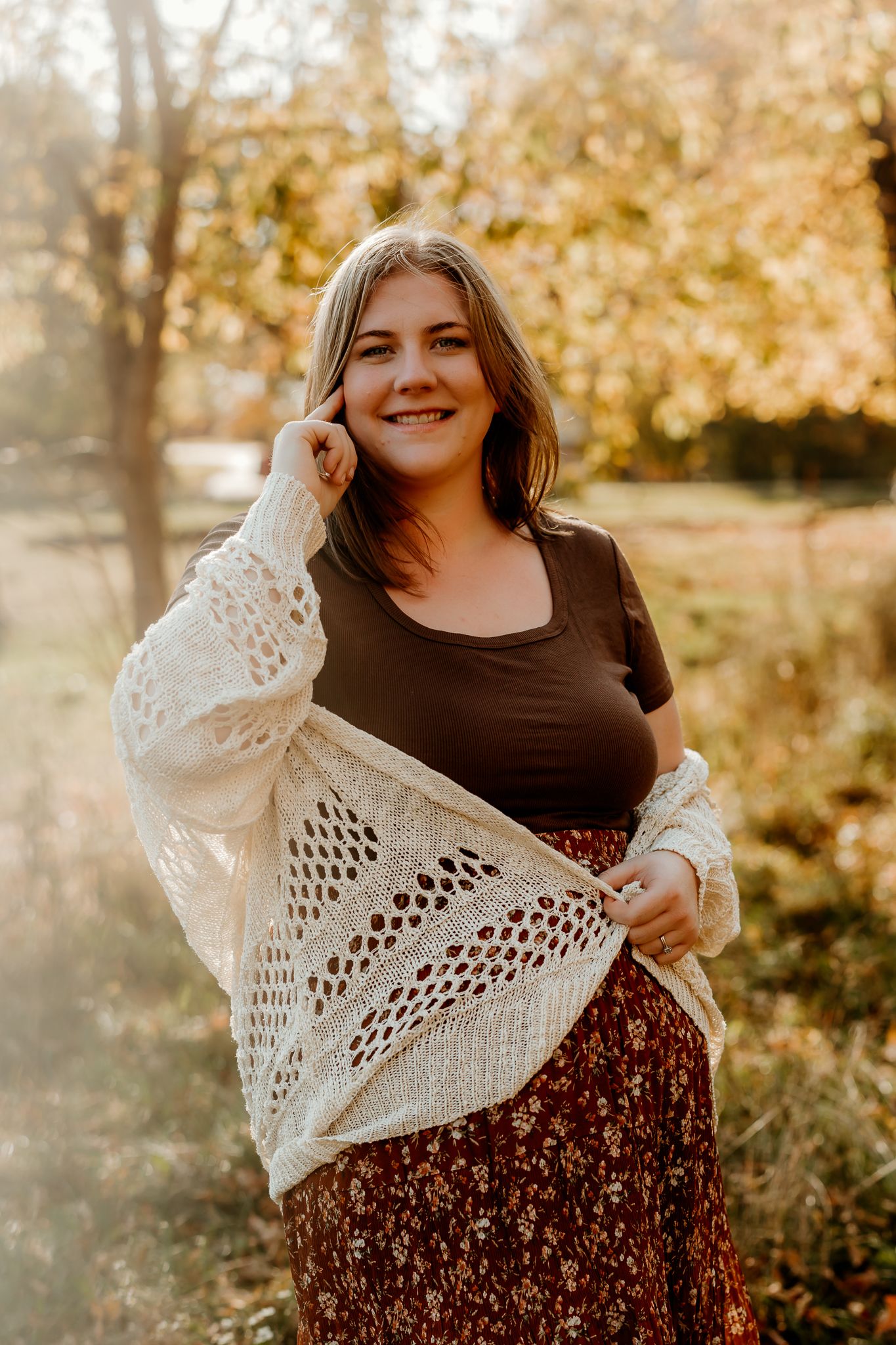 A smiling young woman in a cream knit cardigan and floral skirt poses outdoors among golden autumn trees.