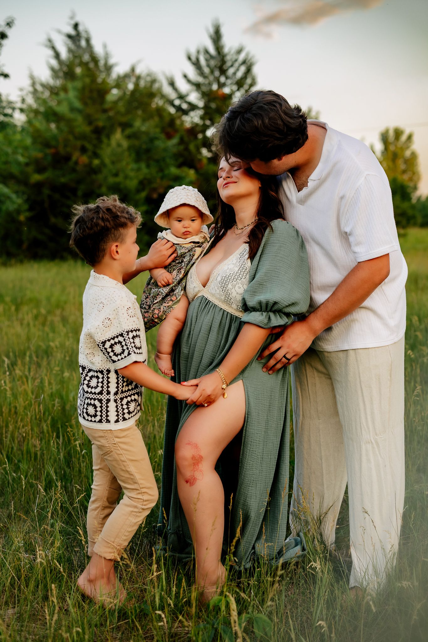 Loving family maternity session outdoors with pregnant mother in green dress, father kissing her head, and two young kids.