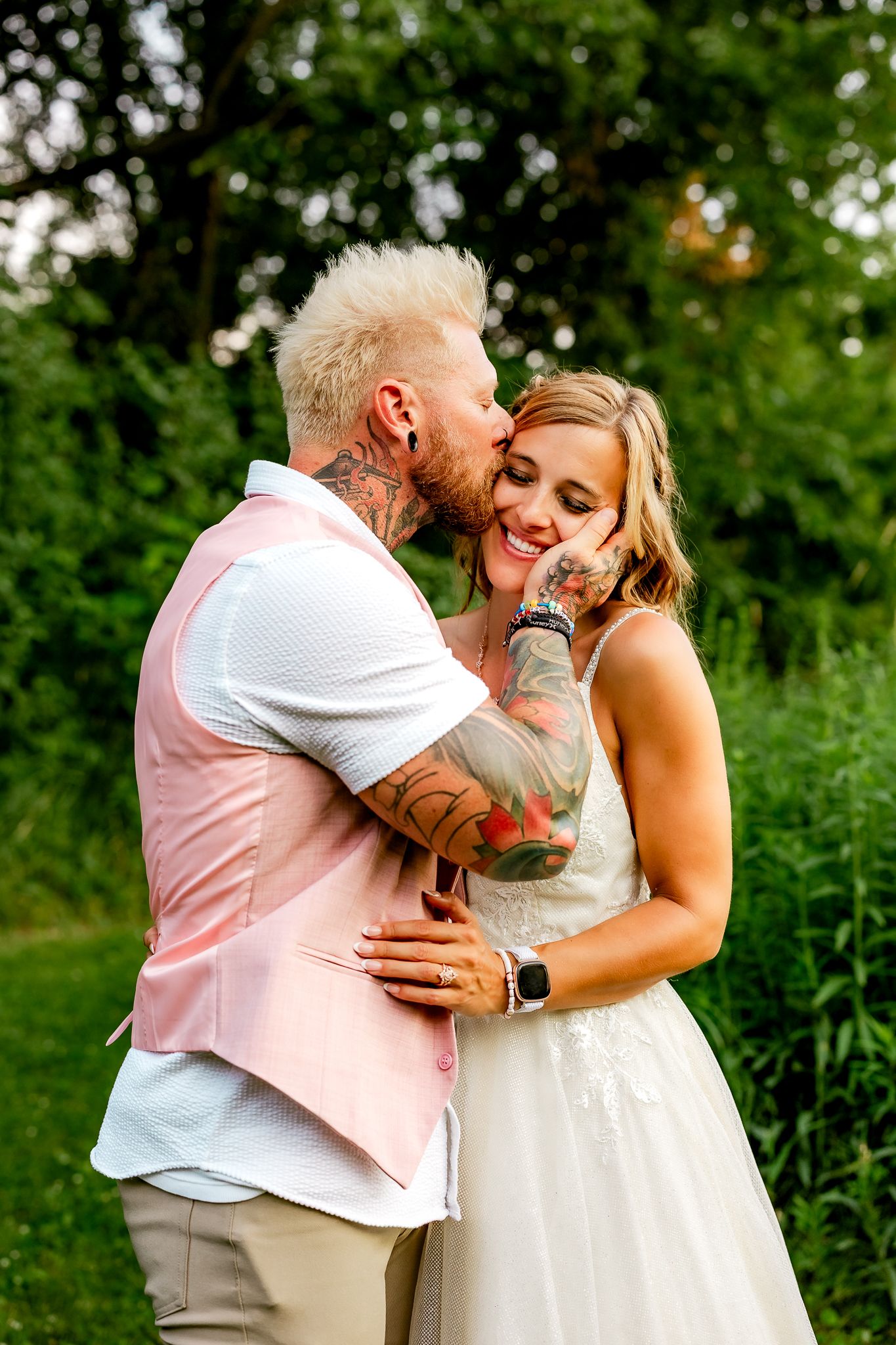 Tattooed groom in pink vest kisses smiling bride in white dress outdoors amid lush green trees.
