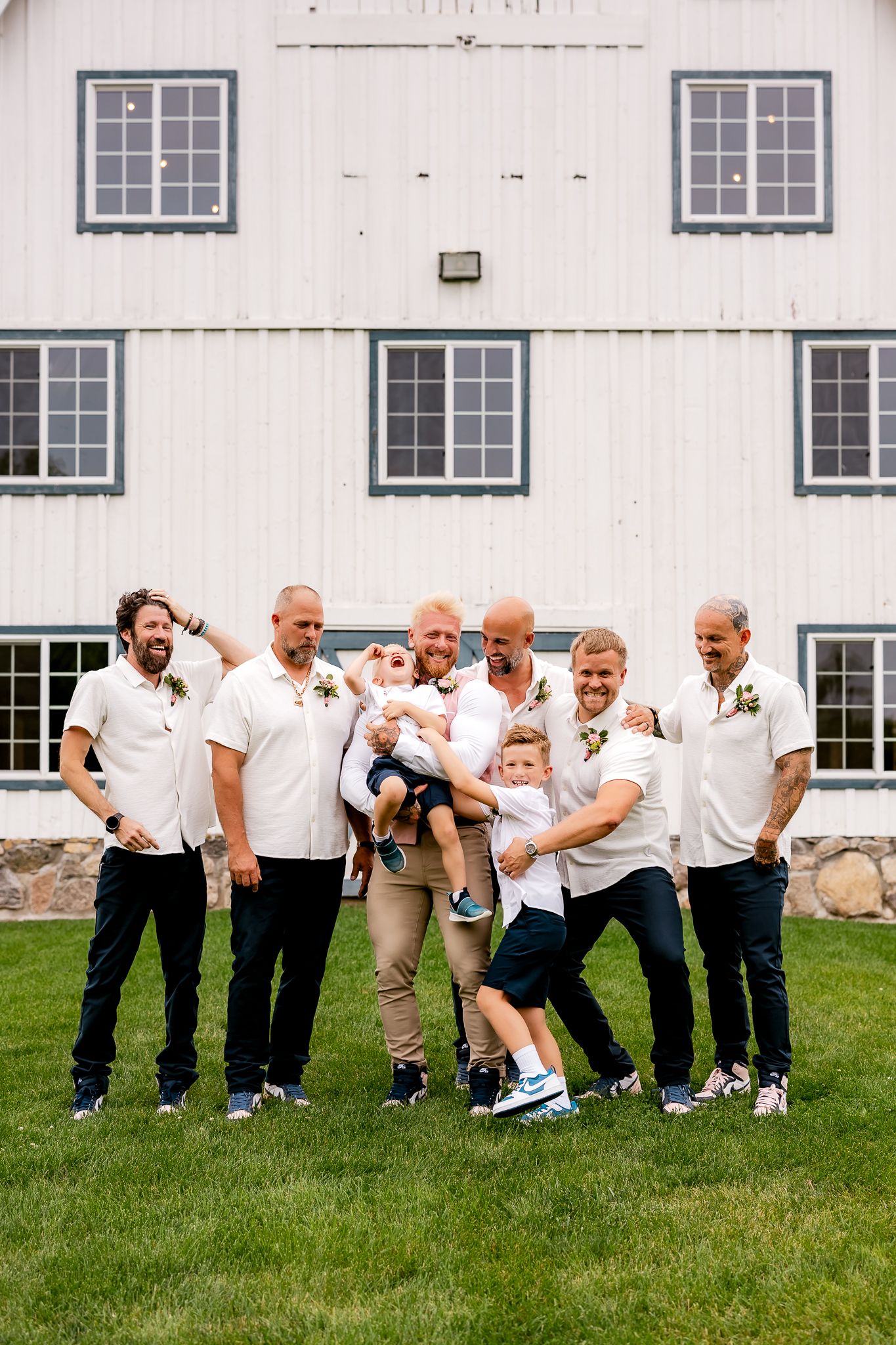 Groomsmen in white polo shirts laugh and pose with two young children in front of a white barn at a wedding.