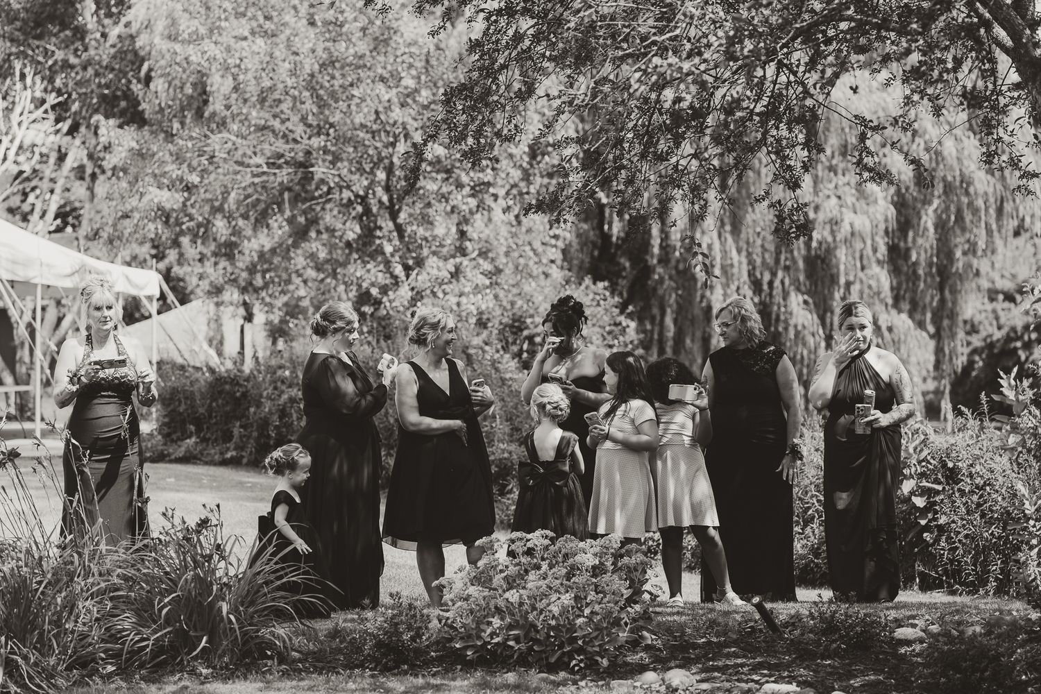 Black and white photo of a group of women in vintage dresses gathered outdoors among lush trees and gardens.