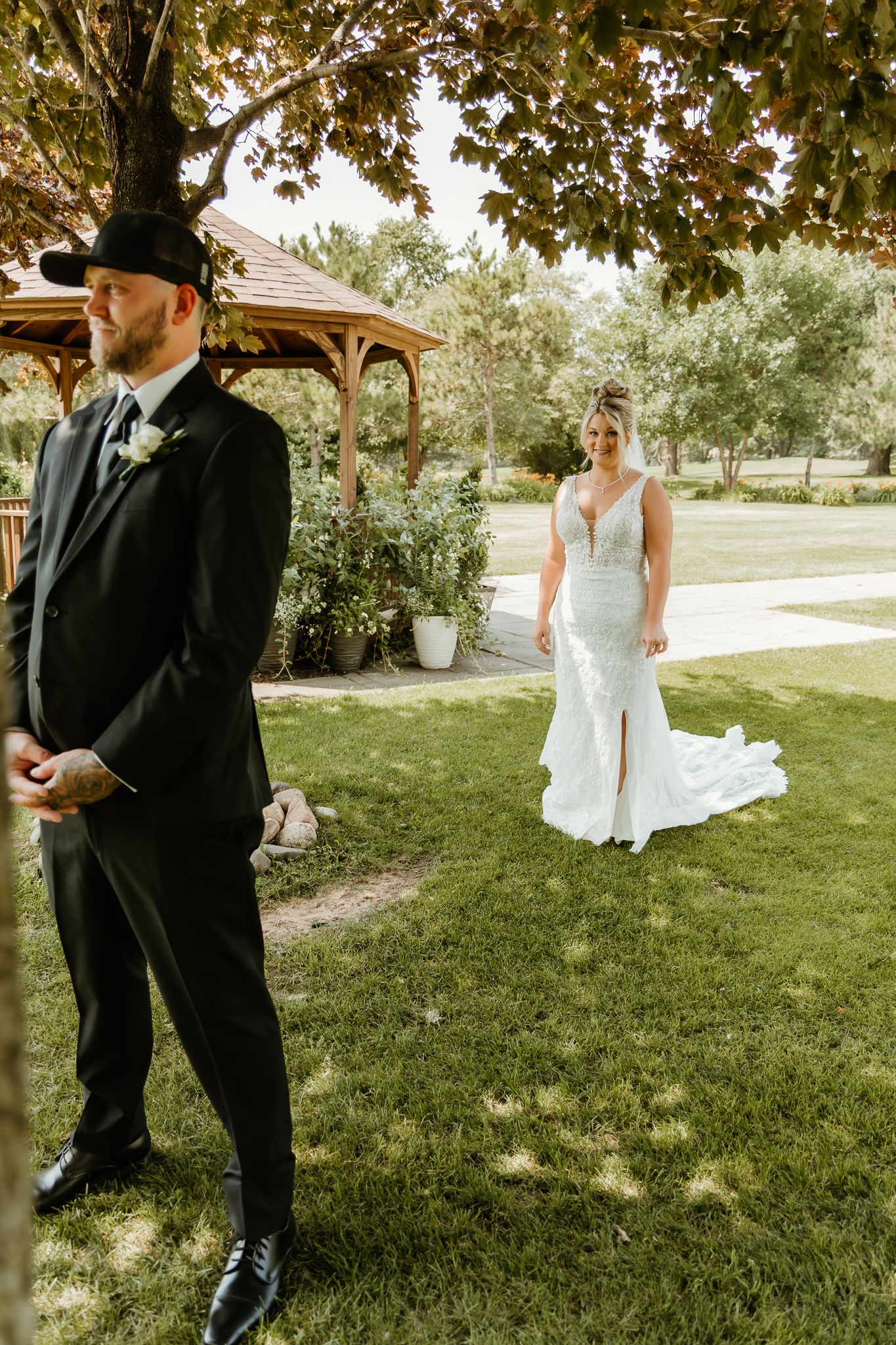 Bride in white lace gown approaches groom in black tuxedo during outdoor first look at garden wedding venue.
