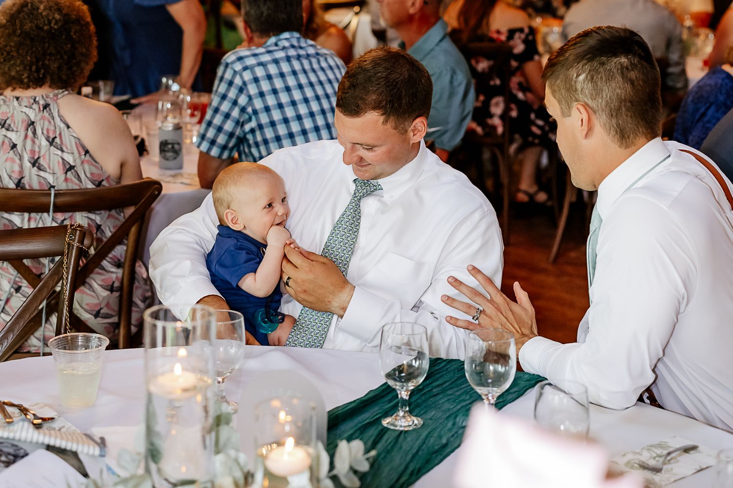 Two men in dress shirts interact with a baby in a navy suit at an elegant wedding reception dinner table.