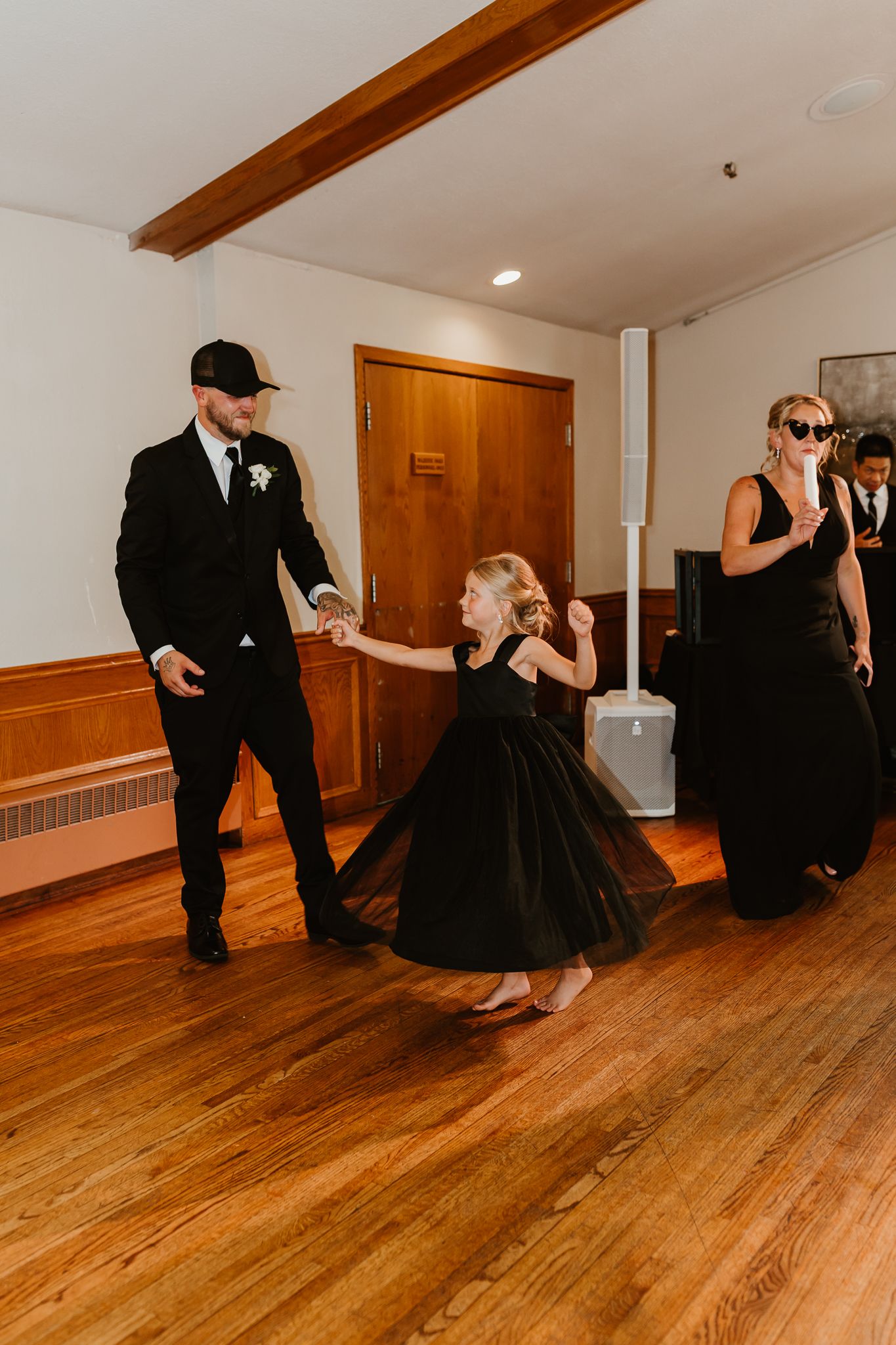 Man in black suit twirls young girl in black dress on wooden dance floor at wedding reception.