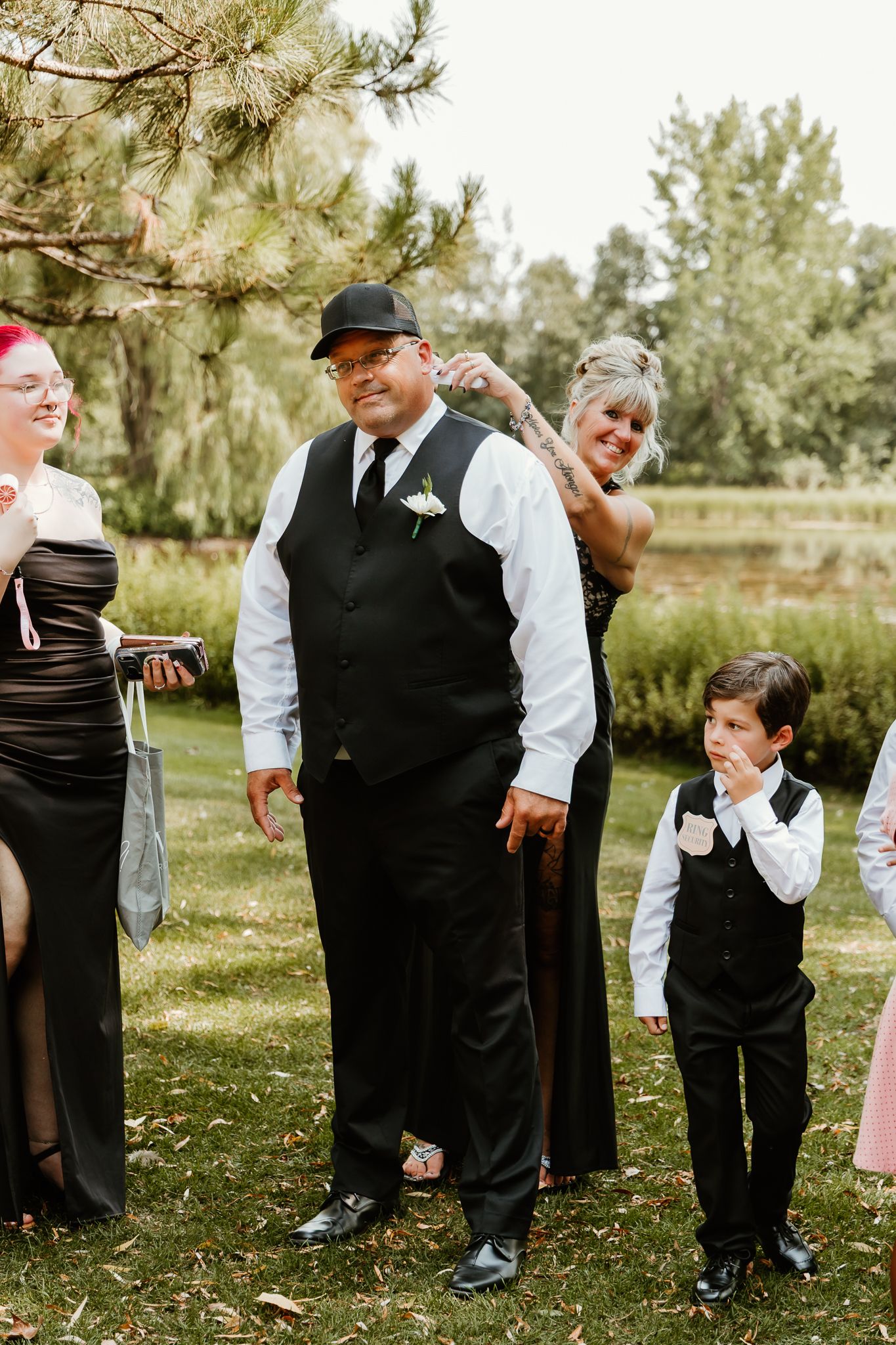 A man in a black vest and cap stands with a young boy in matching attire at an outdoor wedding ceremony.