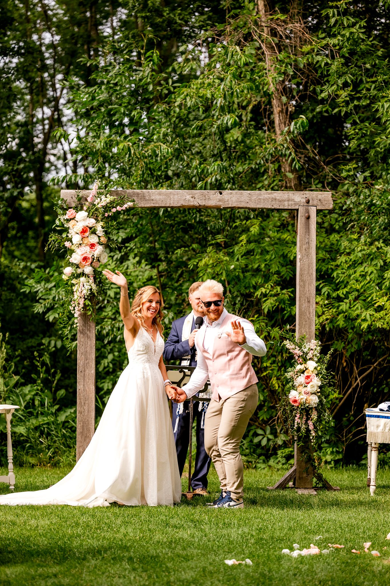 Bride and groom celebrate under a floral wooden arch at an outdoor wedding ceremony on a sunny day.