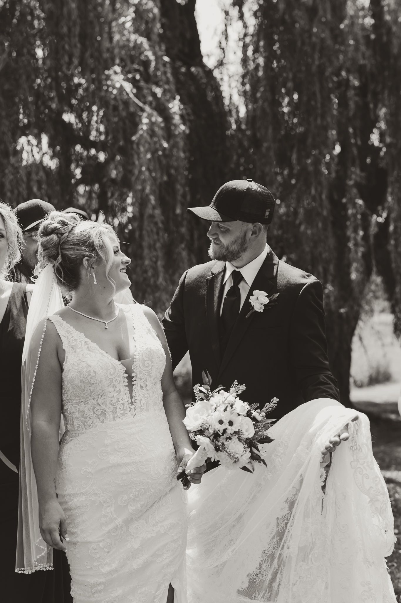 Black and white wedding photo of smiling bride in lace gown and groom in cap holding bouquet under willow tree.