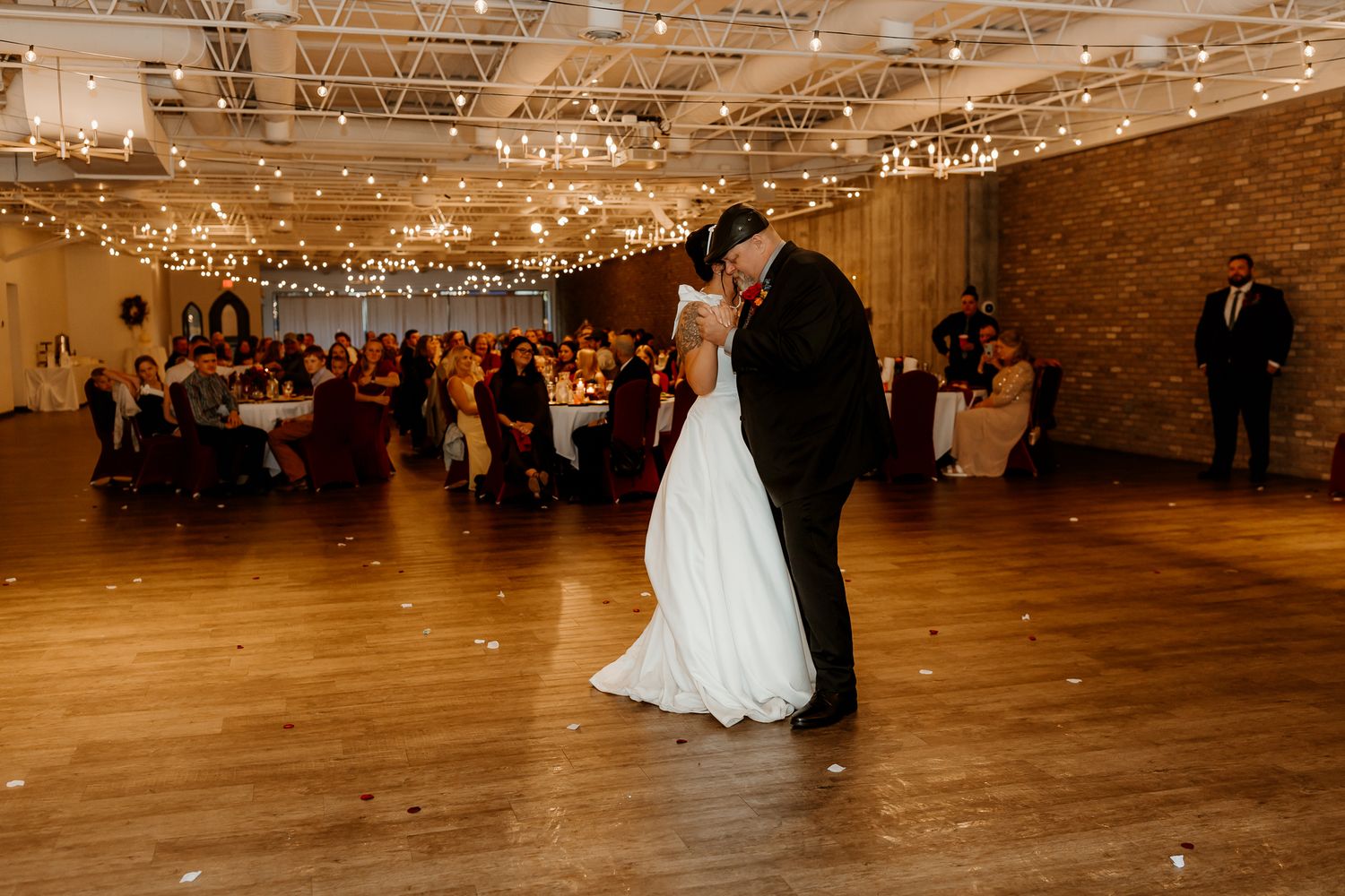 Bride in a white gown and groom in a dark suit share a first dance on a wooden floor beneath string lights as wedding guests watch.
