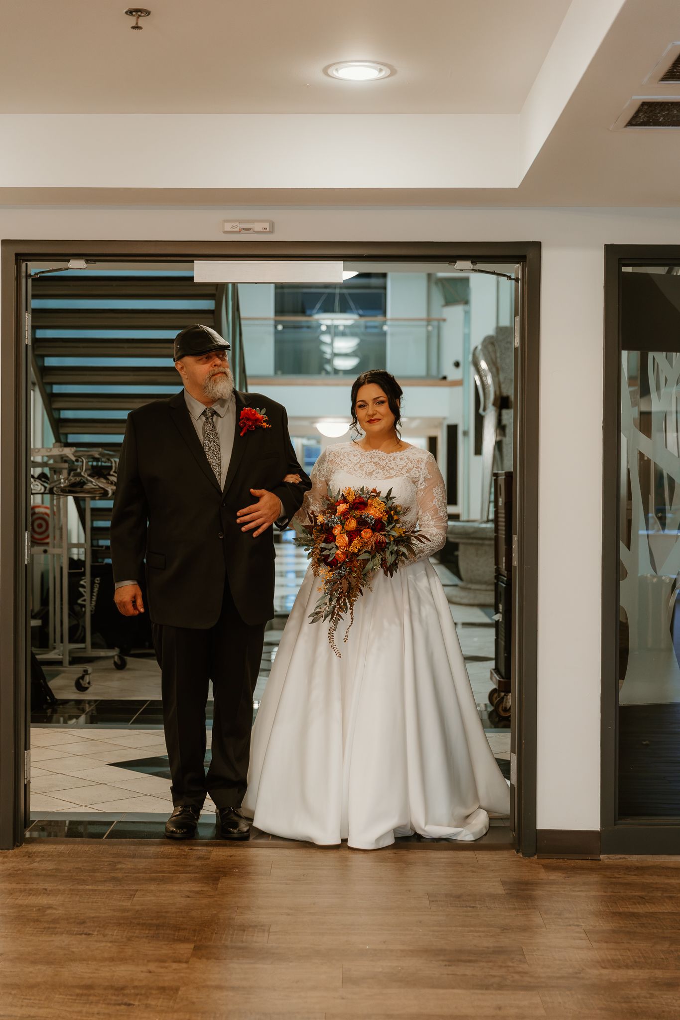 A bride in a white lace gown holds an autumn bouquet while standing arm in arm with a bearded man in a dark suit in a doorway.