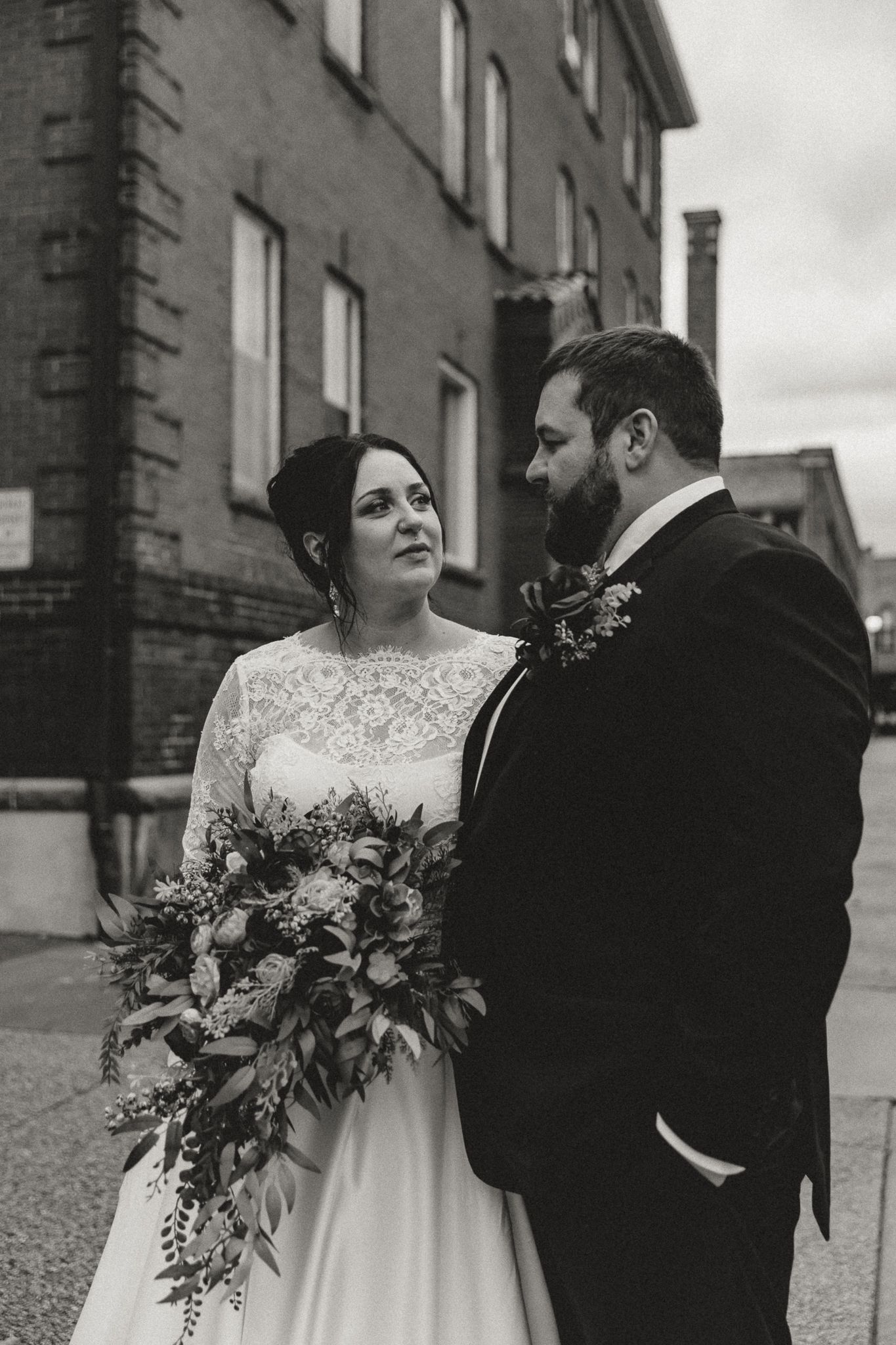 Black and white photo of a bride in a lace gown holding a floral bouquet beside a bearded groom in a dark suit near a brick building.