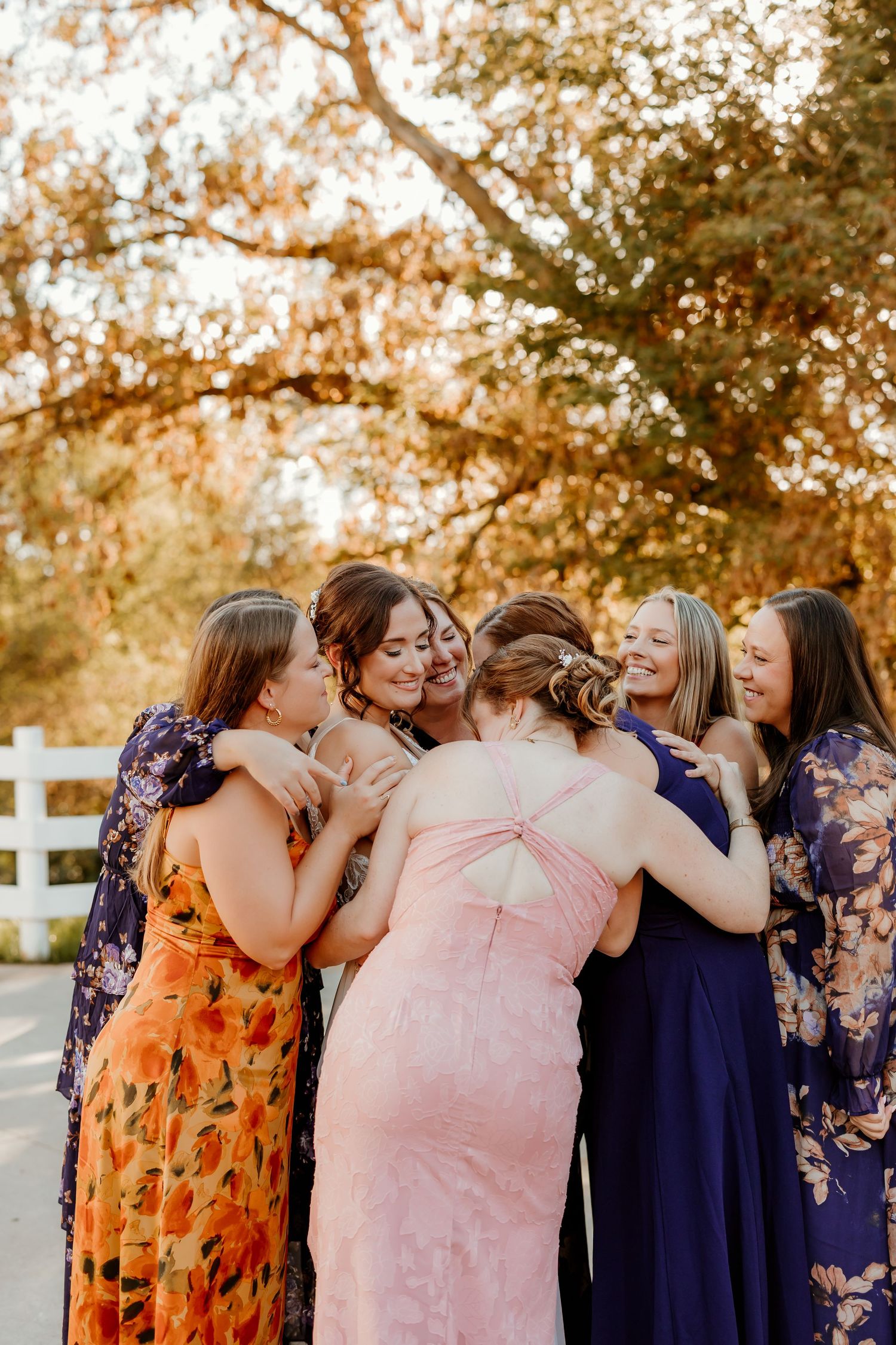 Group of women in colorful floral and pastel dresses share a joyful group hug outdoors beneath autumn trees near a white fence.