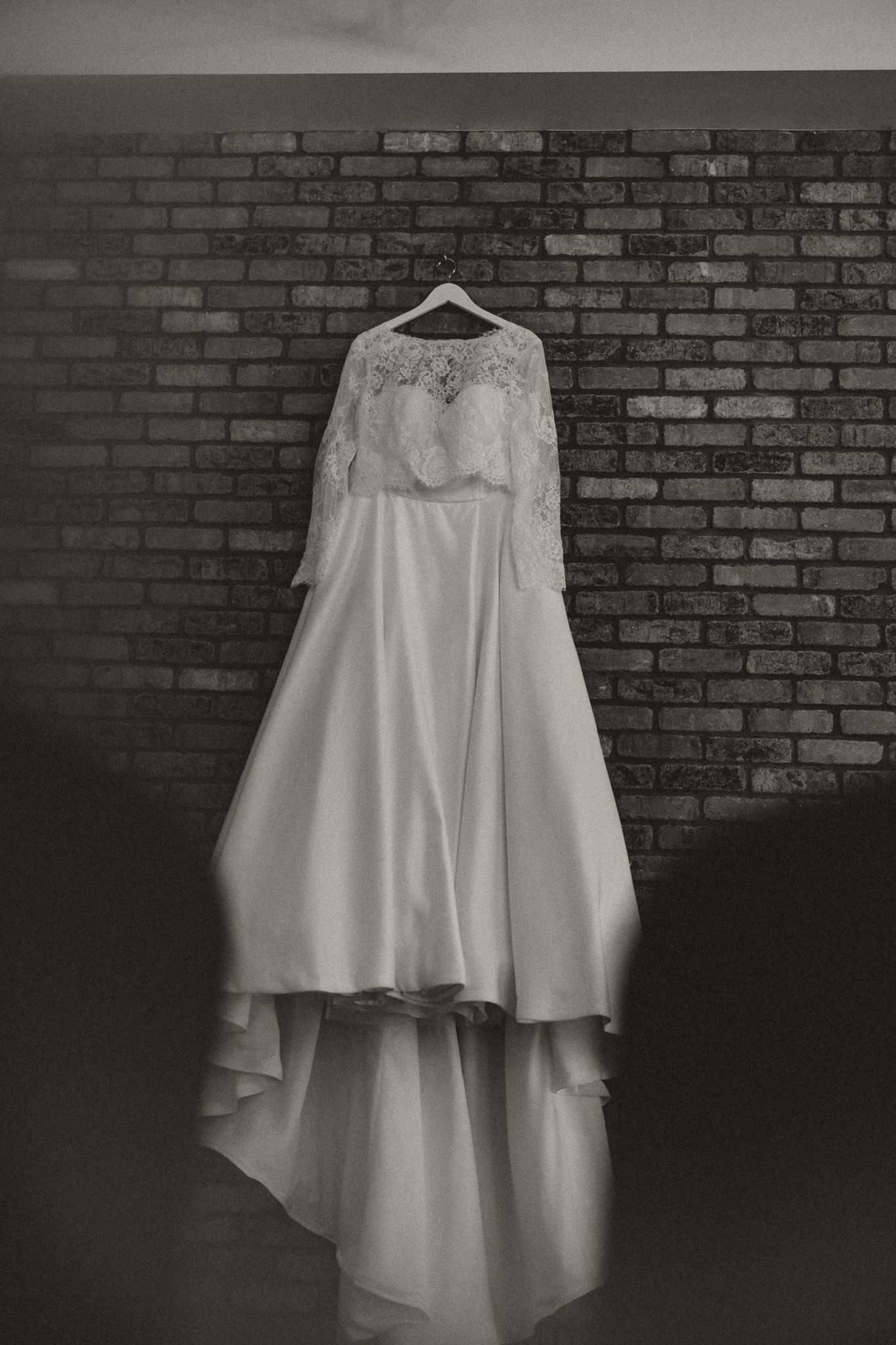 Black and white photo of a lace-sleeved wedding dress with a flowing satin skirt and train hanging on a brick wall.