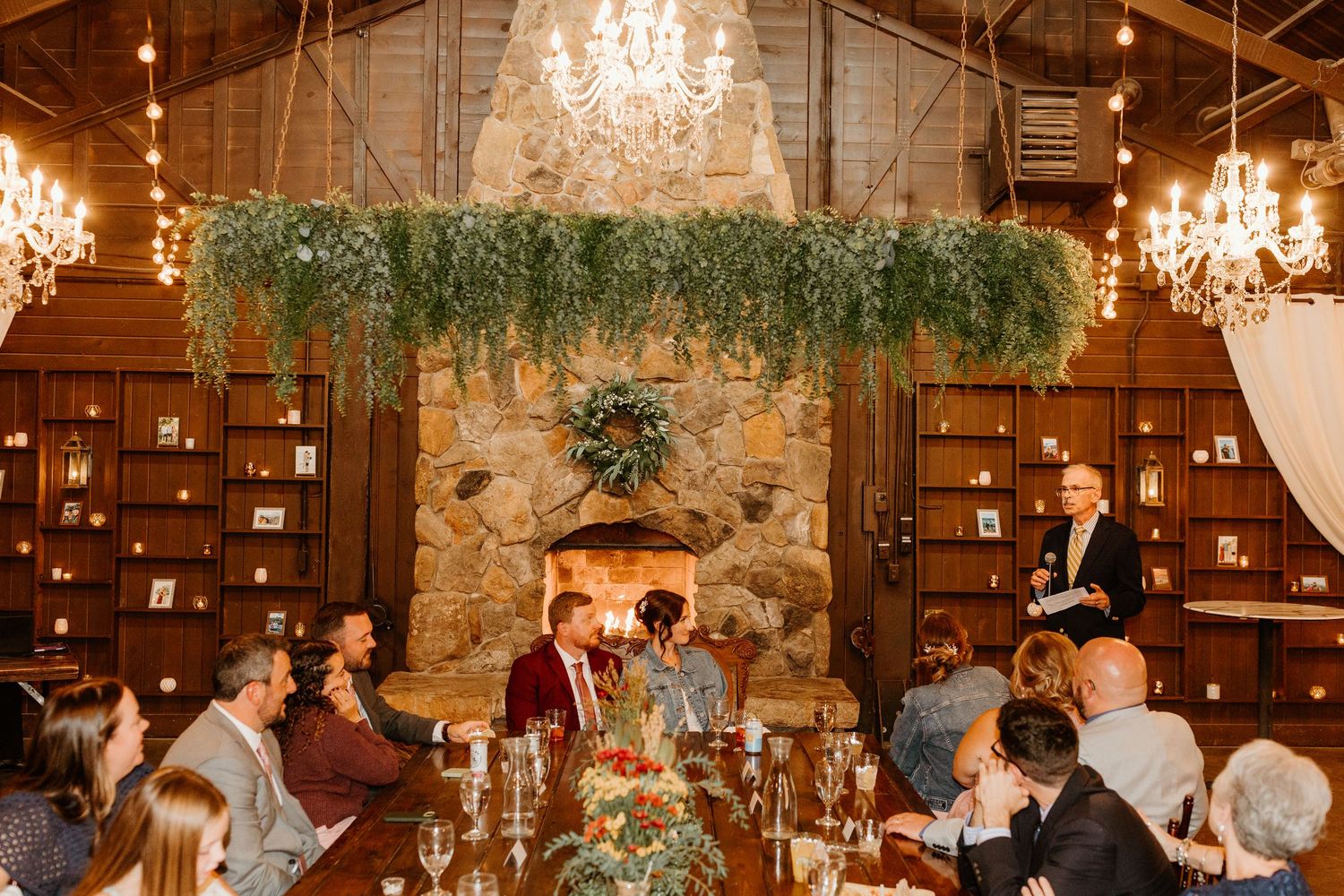 Wedding guests seated at long tables listen to a speaker in a rustic lodge with a stone fireplace, hanging greenery and crystal chandeliers.