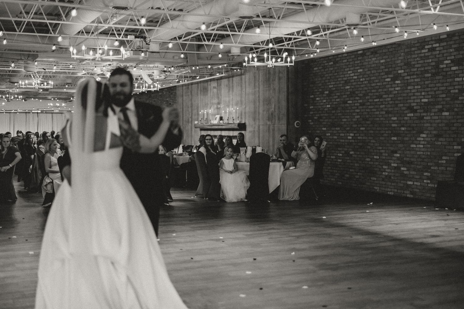 Bride and groom share a first dance in a rustic brick venue while wedding guests watch from tables in this black and white photo.