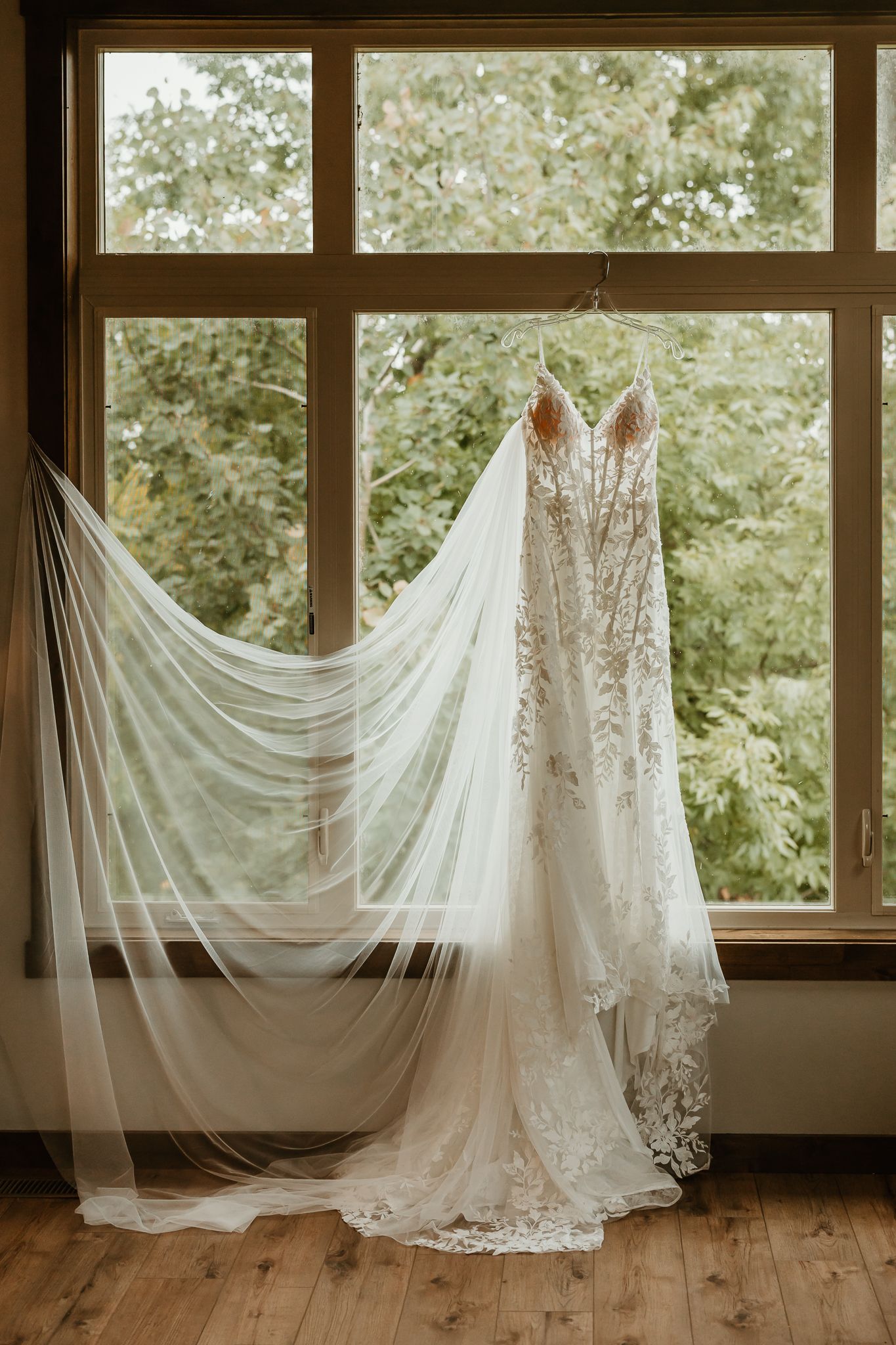 Lace wedding dress with a long sheer veil hanging in front of a large window overlooking green trees, above a wood plank floor.
