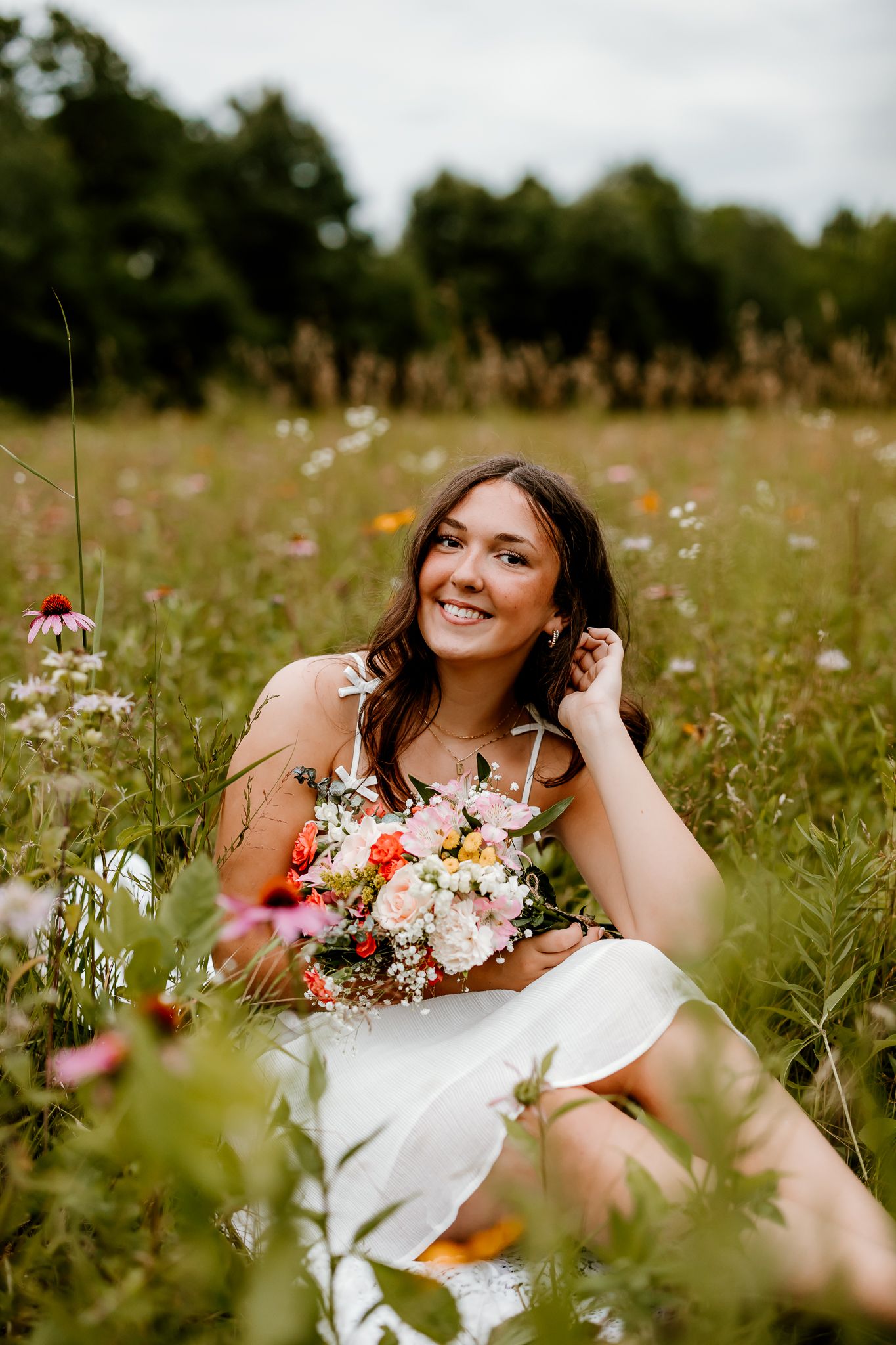 Smiling bride in white dress sitting in wildflower meadow holding colorful bouquet with trees in background.