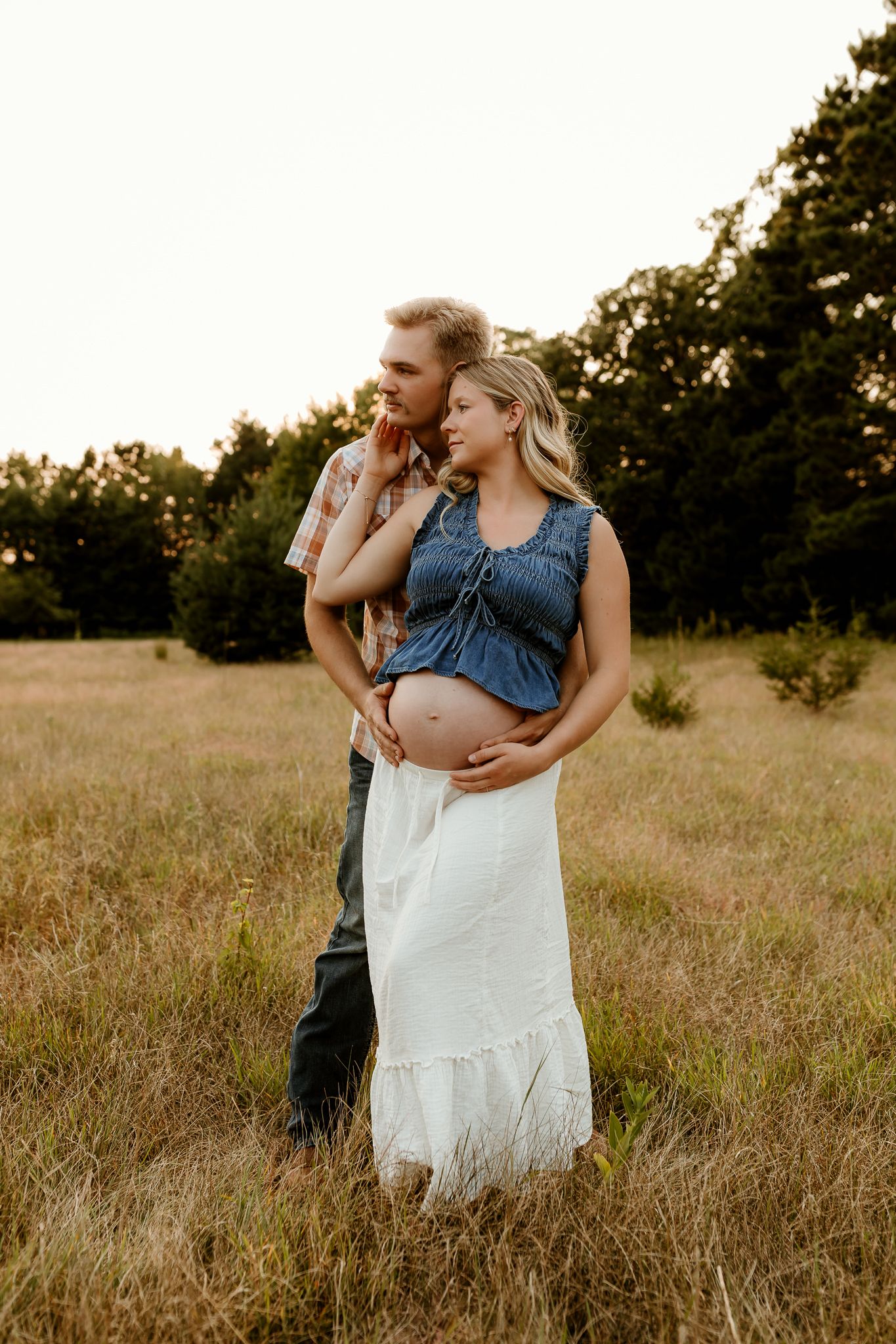 Couple in golden field, man embracing pregnant woman in blue top and white skirt, maternity photo session at sunset.