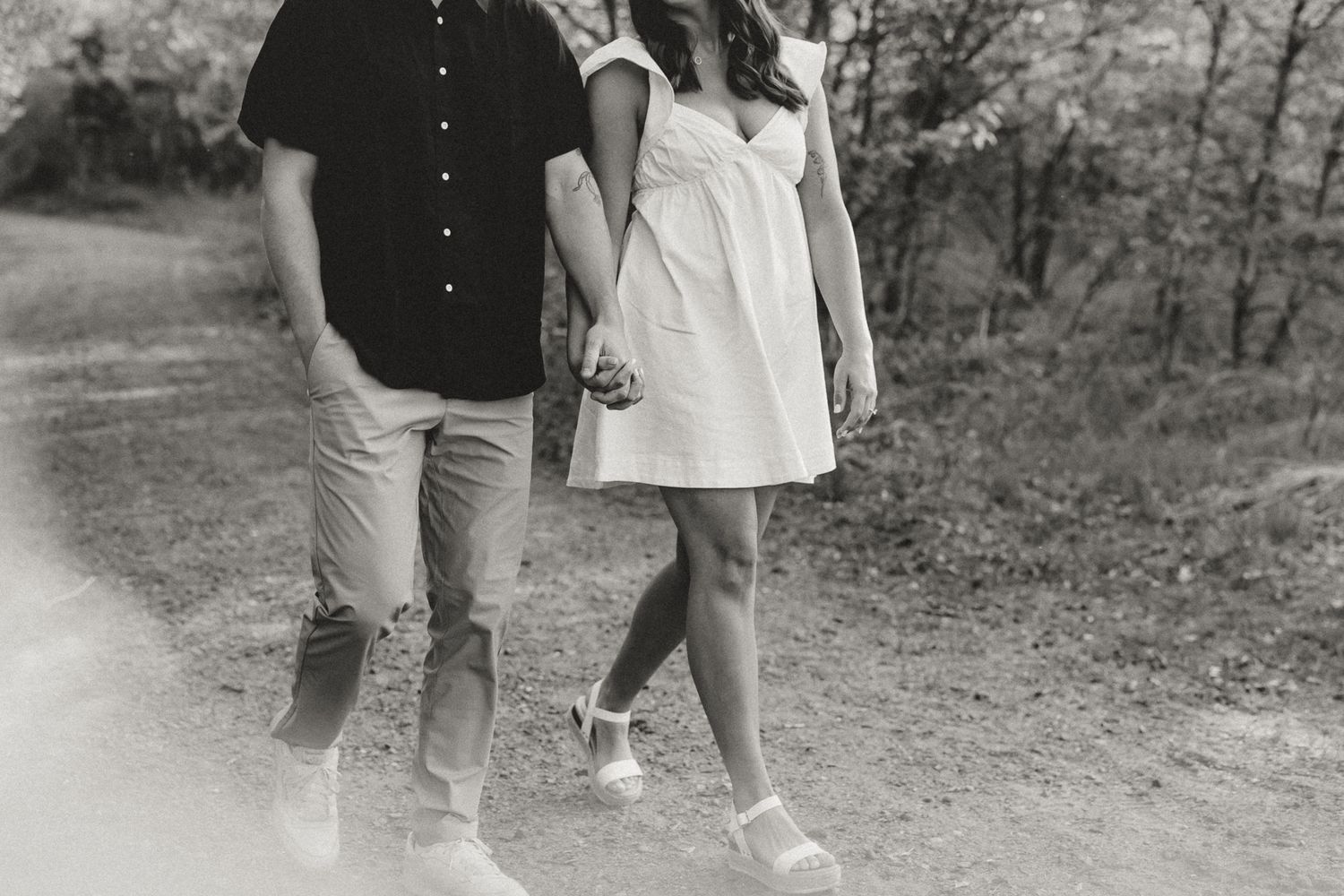 Black and white photo of a couple holding hands while walking on a dirt path surrounded by trees.